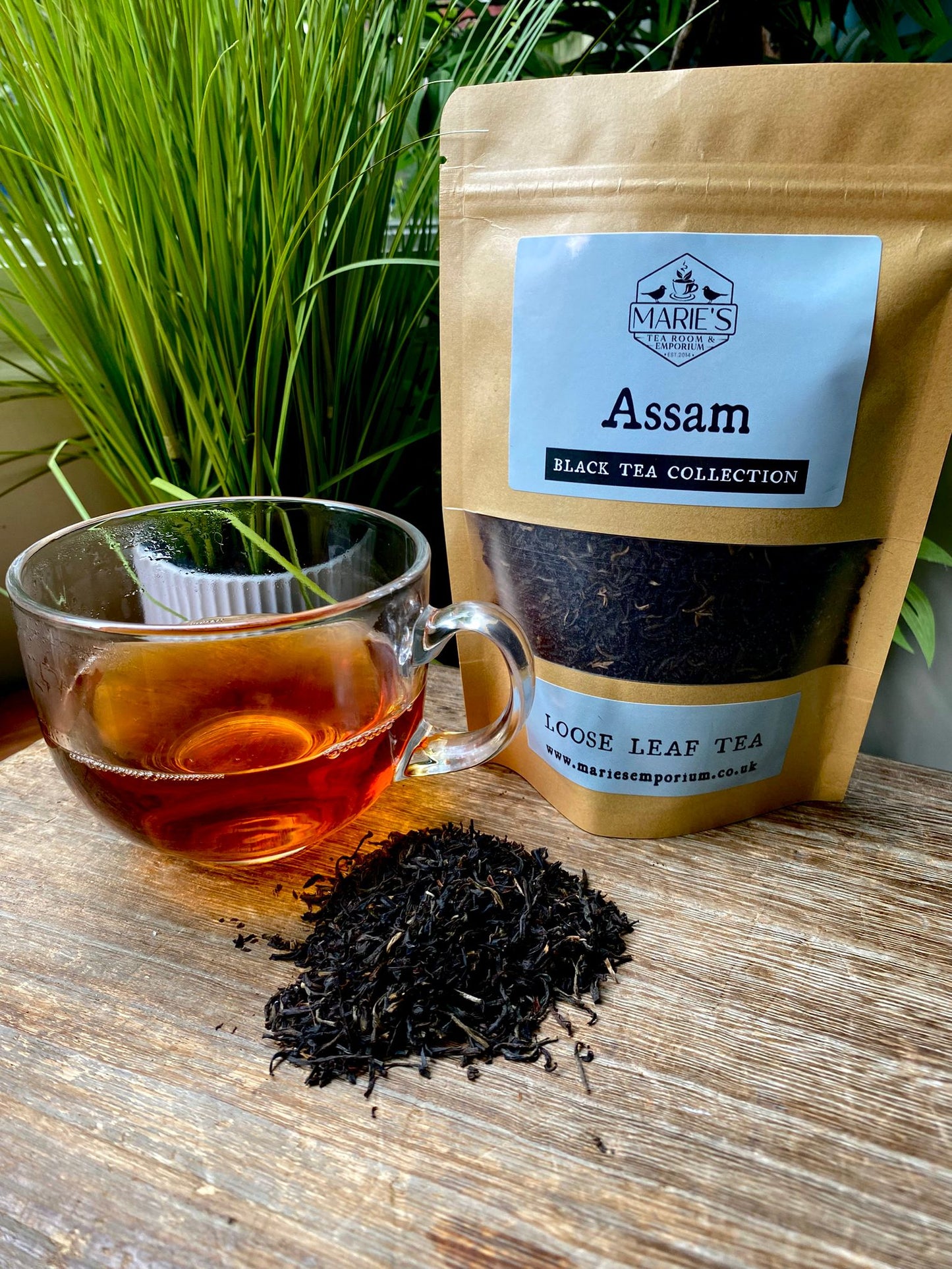 A clear glass cup of Assam tea sits next to a pile of loose leaf tea and a brown package labeled "Assam Loose Leaf Tea" on a wooden surface, with green plants in the background.