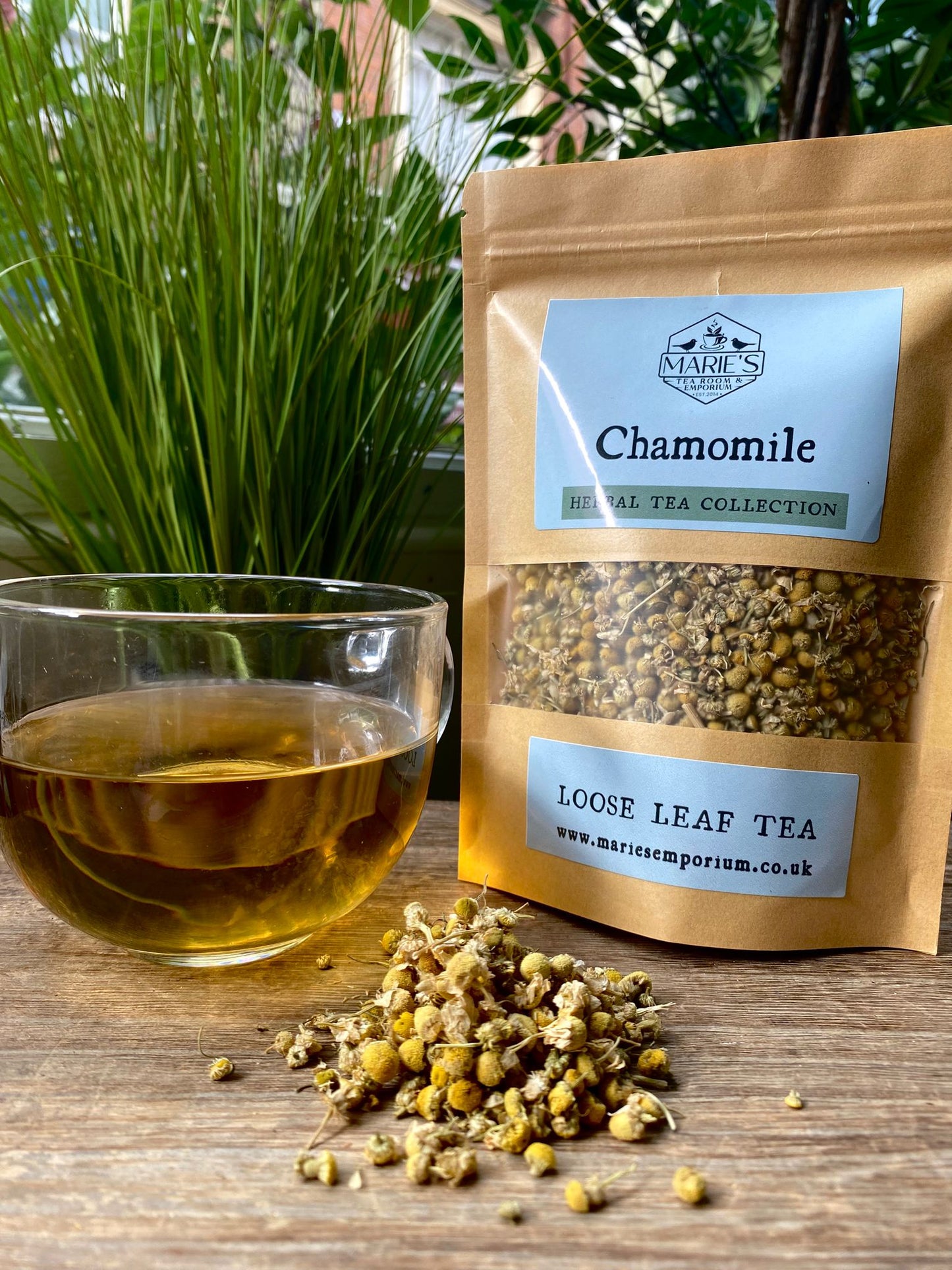 marie's Loose leaf chamomile tea package with a glass of brewed tea and scattered chamomile flowers on a wooden surface.