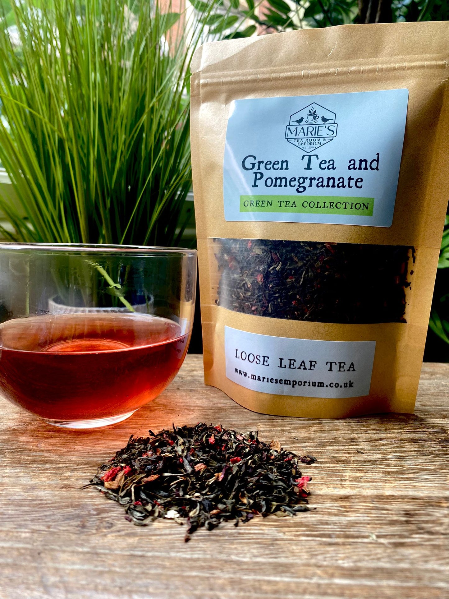 marie's Green Tea and Pomegranate tea package with a glass of tea and loose tea leaves on a wooden surface.