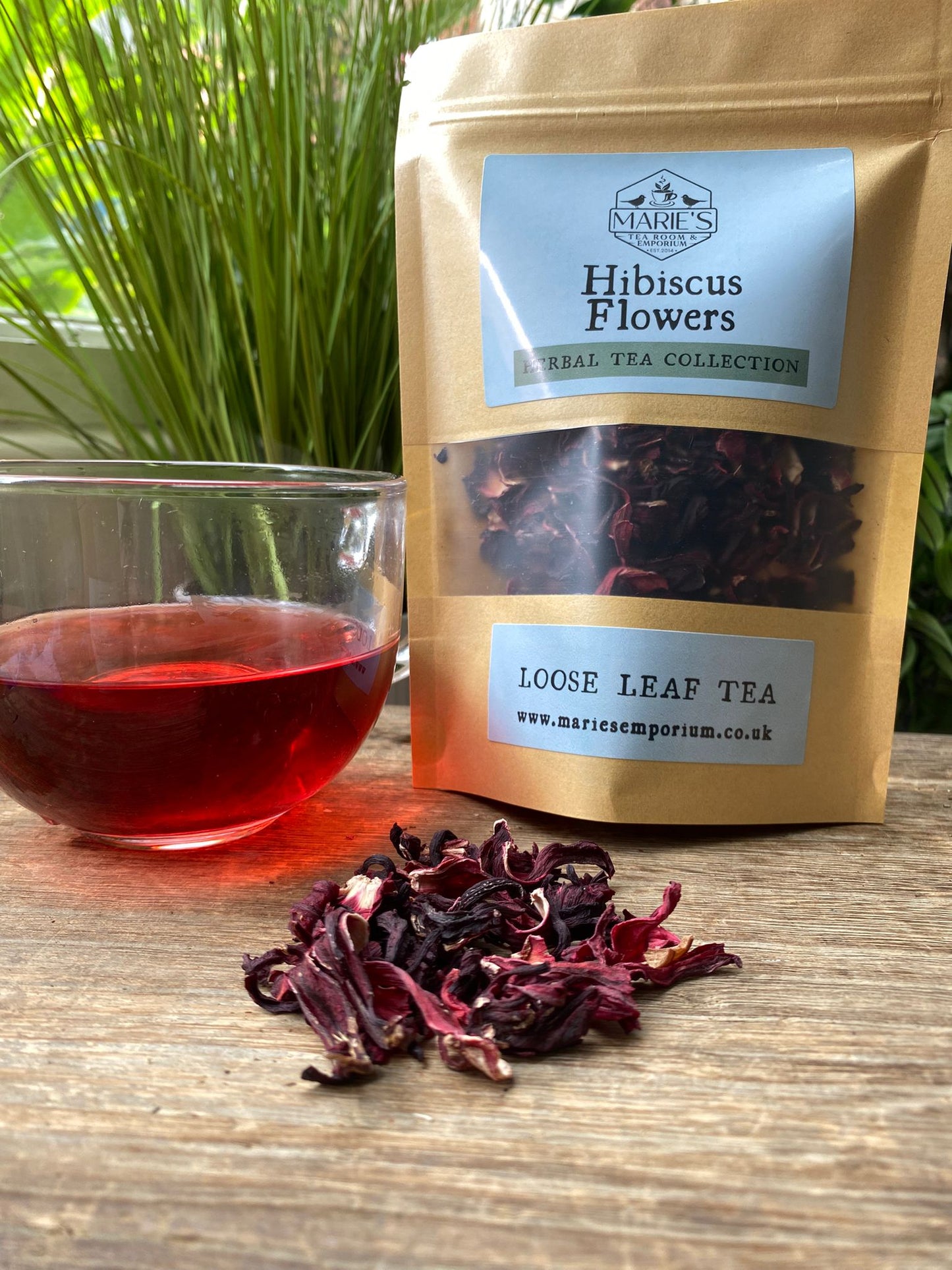 Hibiscus flowers, a glass of brewed tea, and a package labeled 'Hibiscus Flowers Loose Leaf Tea' on a wooden surface.