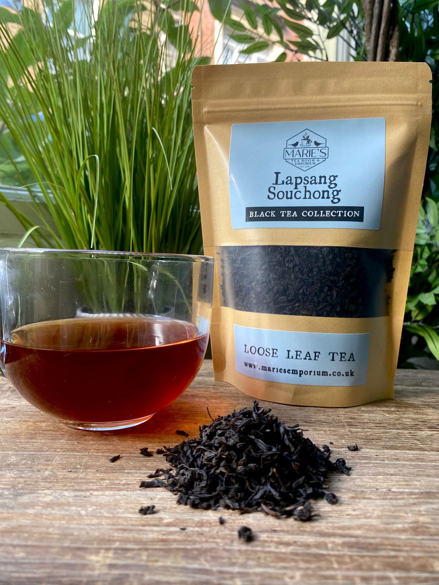Bag of Lapsang Souchong tea, a glass with brewed tea, and loose tea leaves on a wooden surface with plants in the background.