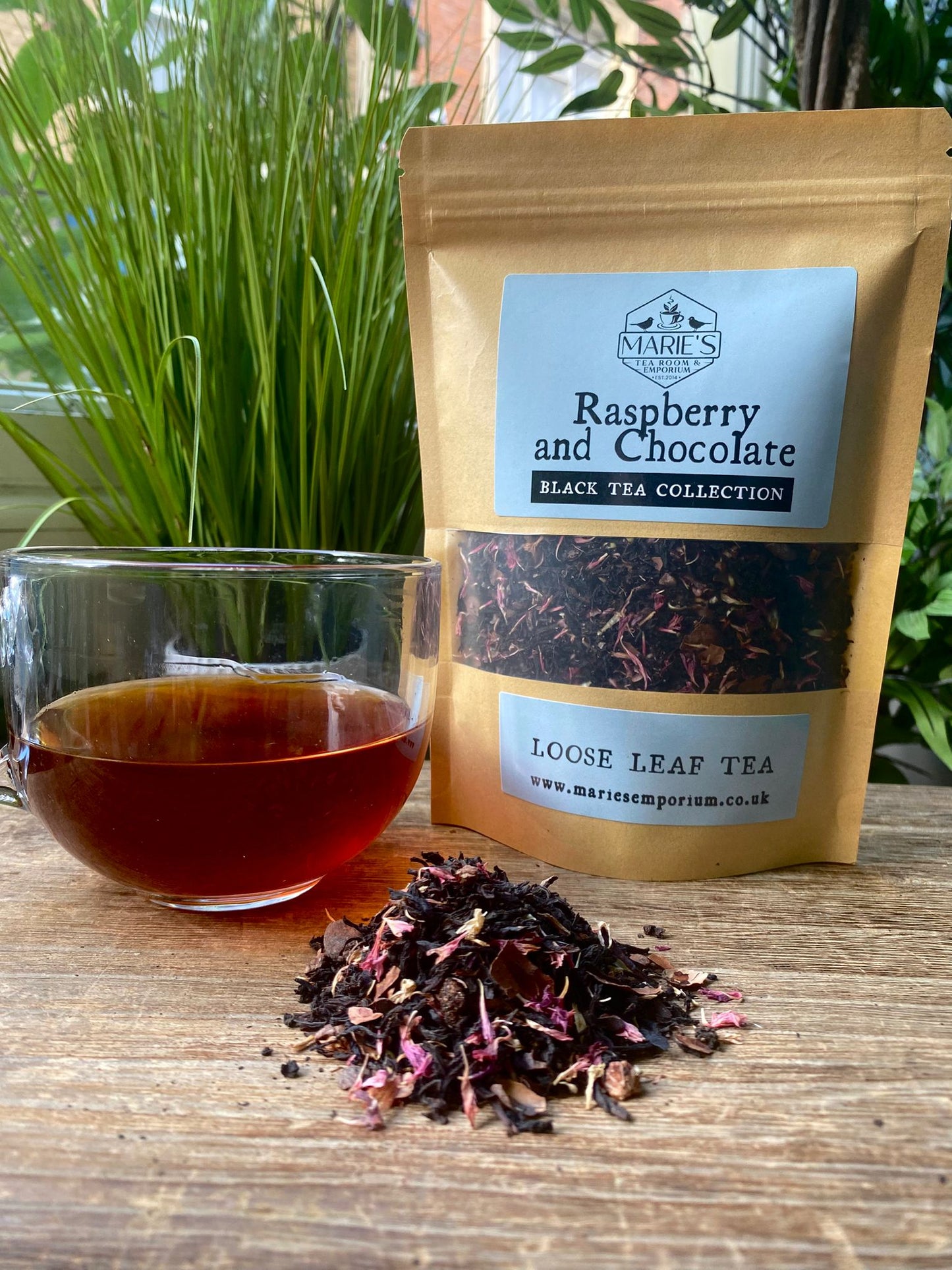 A glass cup with dark raspberry and chocolate tea sits beside a pouch labeled “Maries Raspberry and Chocolate Loose Leaf Tea” on a wooden table with leafy plants in the background.