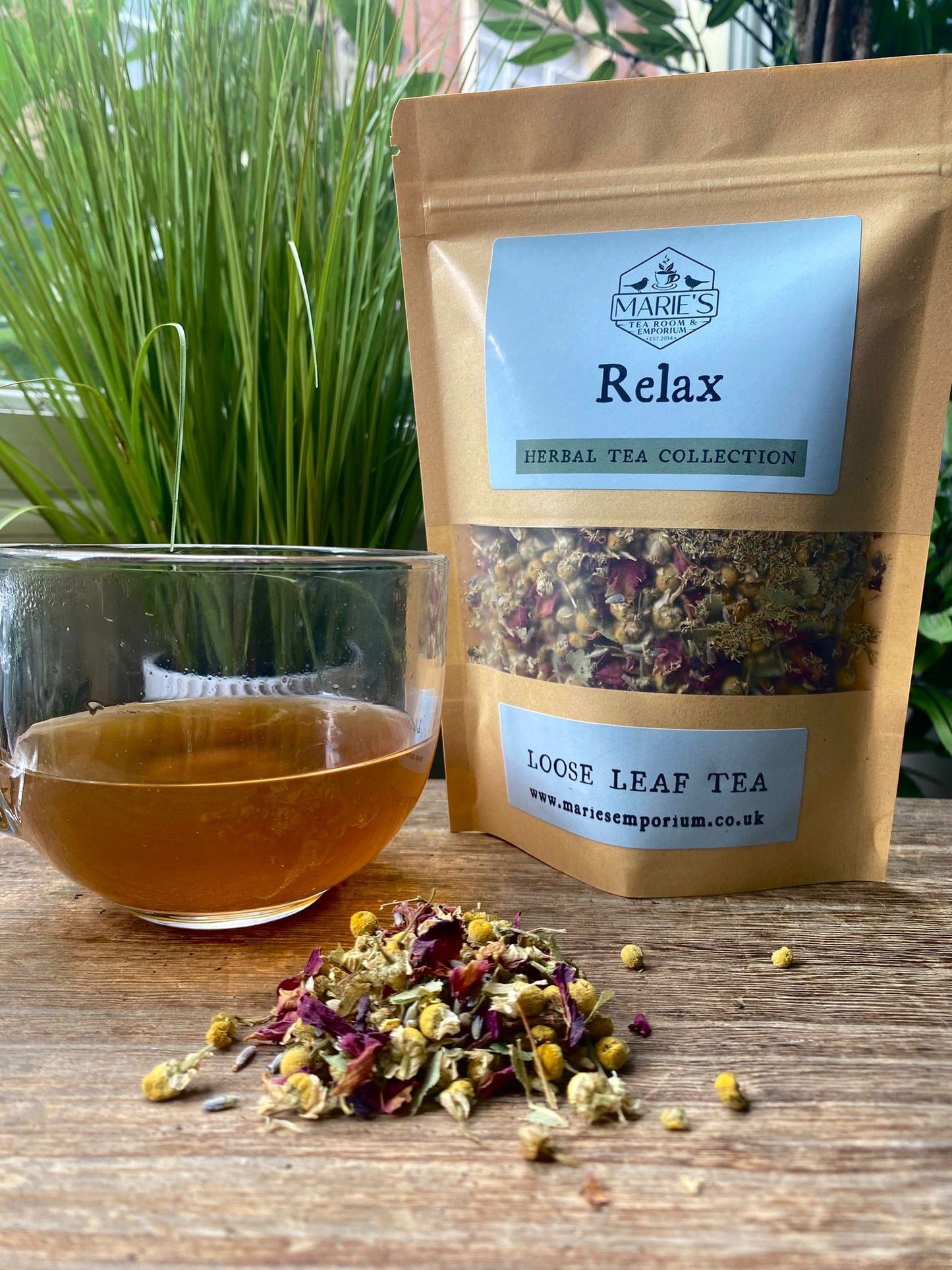 Marie's Emporium herbal tea collection with a bag labeled 'Relax', a glass of tea, and loose leaf tea on a wooden surface.