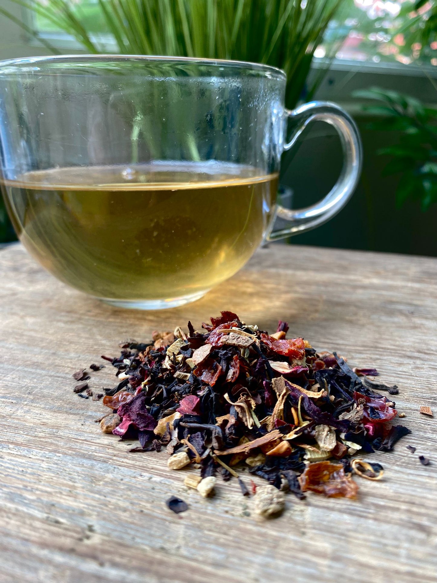 Clear glass mug with Marie's spiced orange ooolog tea and a pile of dried tea leaves on a wooden surface with plants in the background.