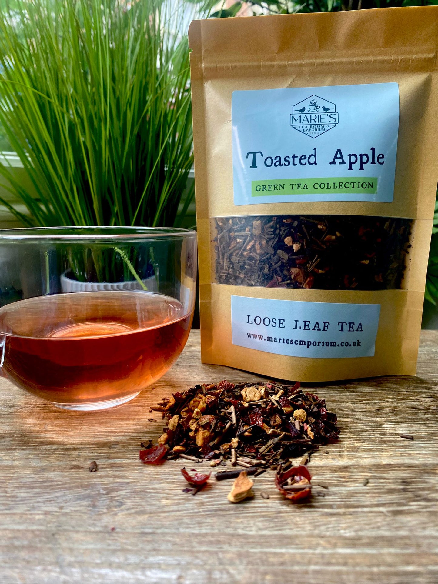 Marie's Emporium Toasted Apple green tea package with a glass of tea and loose tea leaves on a wooden surface.