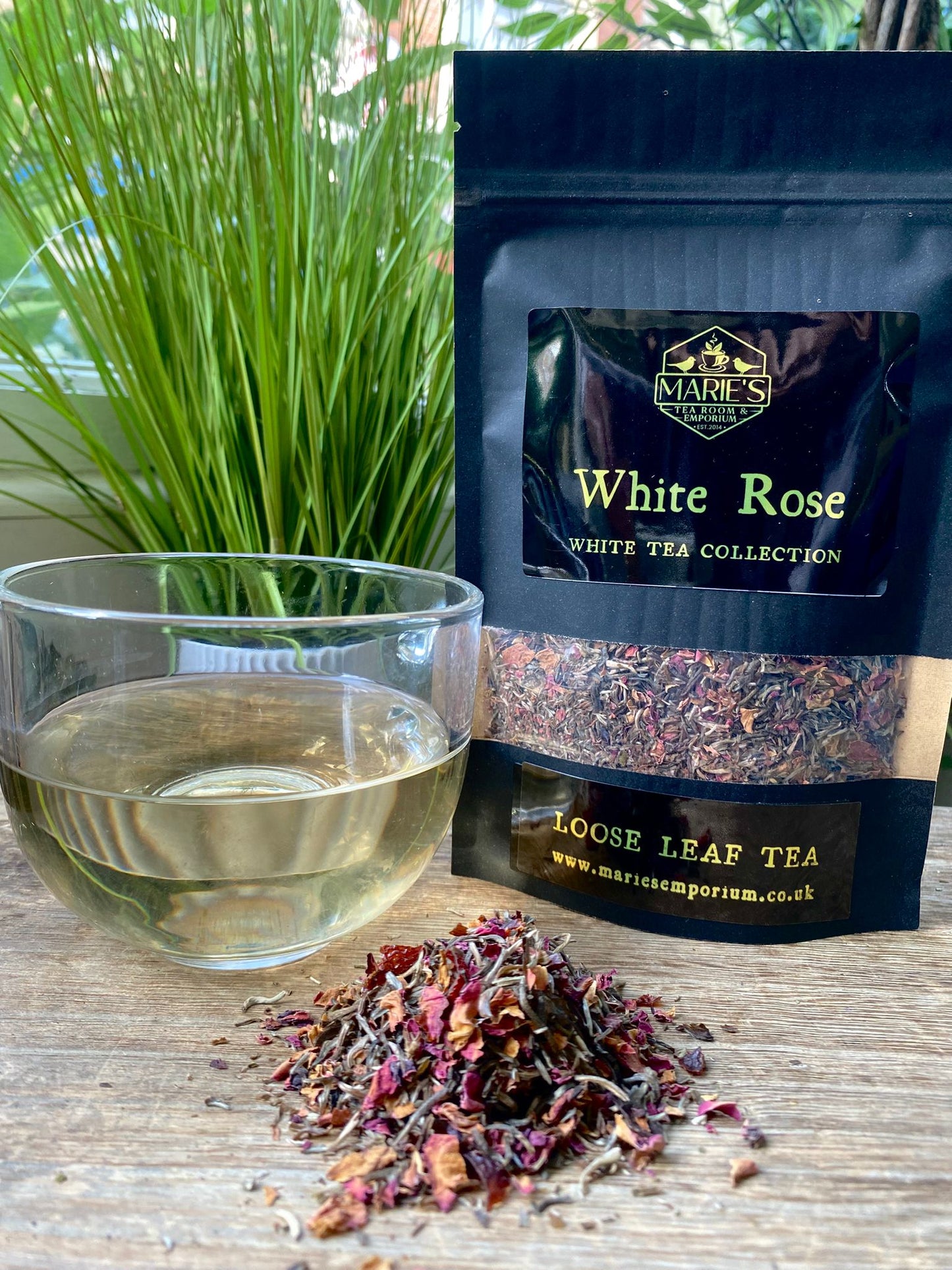 A glass cup of light-colored tea sits on a wooden table next to a package labeled Marie's White Rose loose leaf tea. Dried petals are scattered nearby.