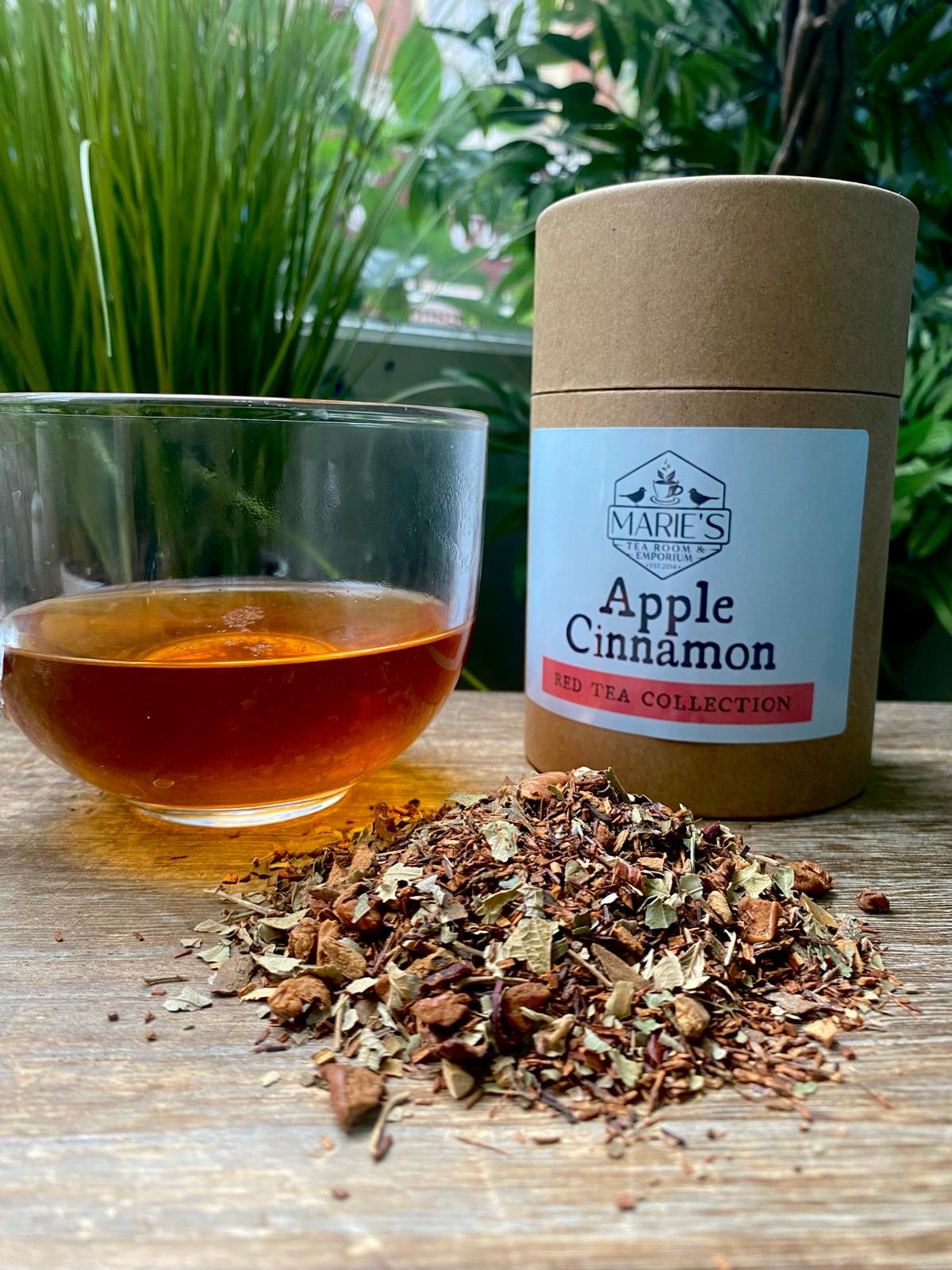 Glass of warm Maries red tea sits on a wooden table next to spilled loose apple cinnamon tea leaves and a labeled brown tea canister. Greenery in the background.