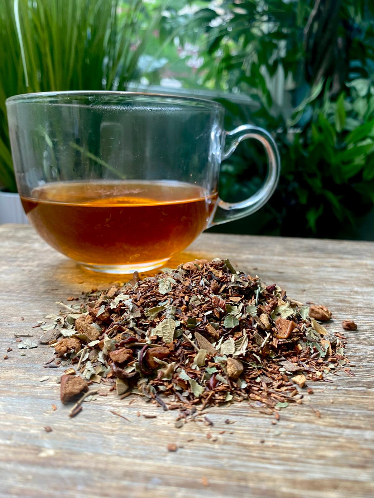 A clear glass teacup with maries Apple Cinnamon tea sits on a wooden table next to a pile of loose leaf tea. Lush green plants in the blurred background.