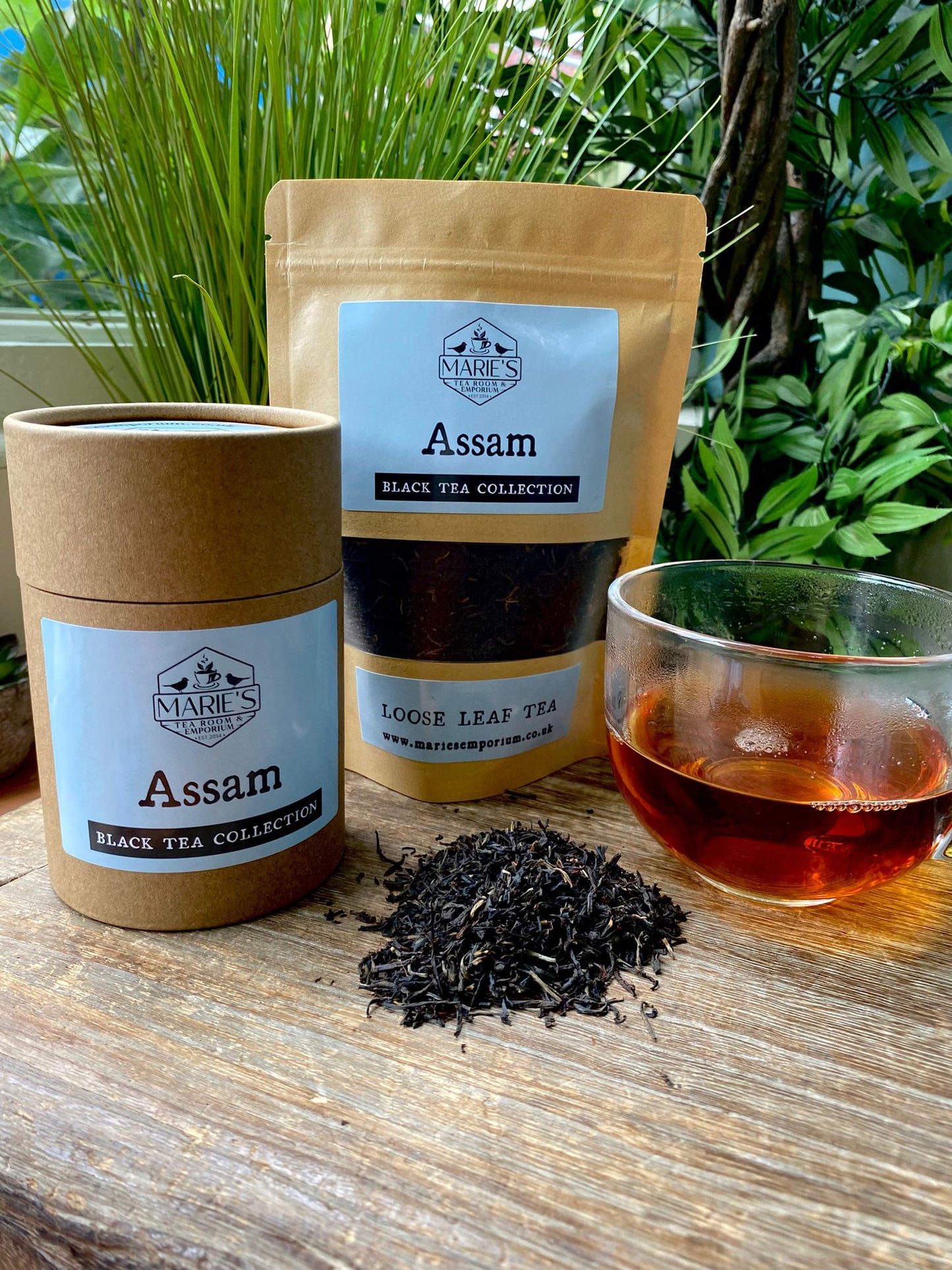 Assam black tea packaging and a cup of tea on a wooden surface with plants in the background.