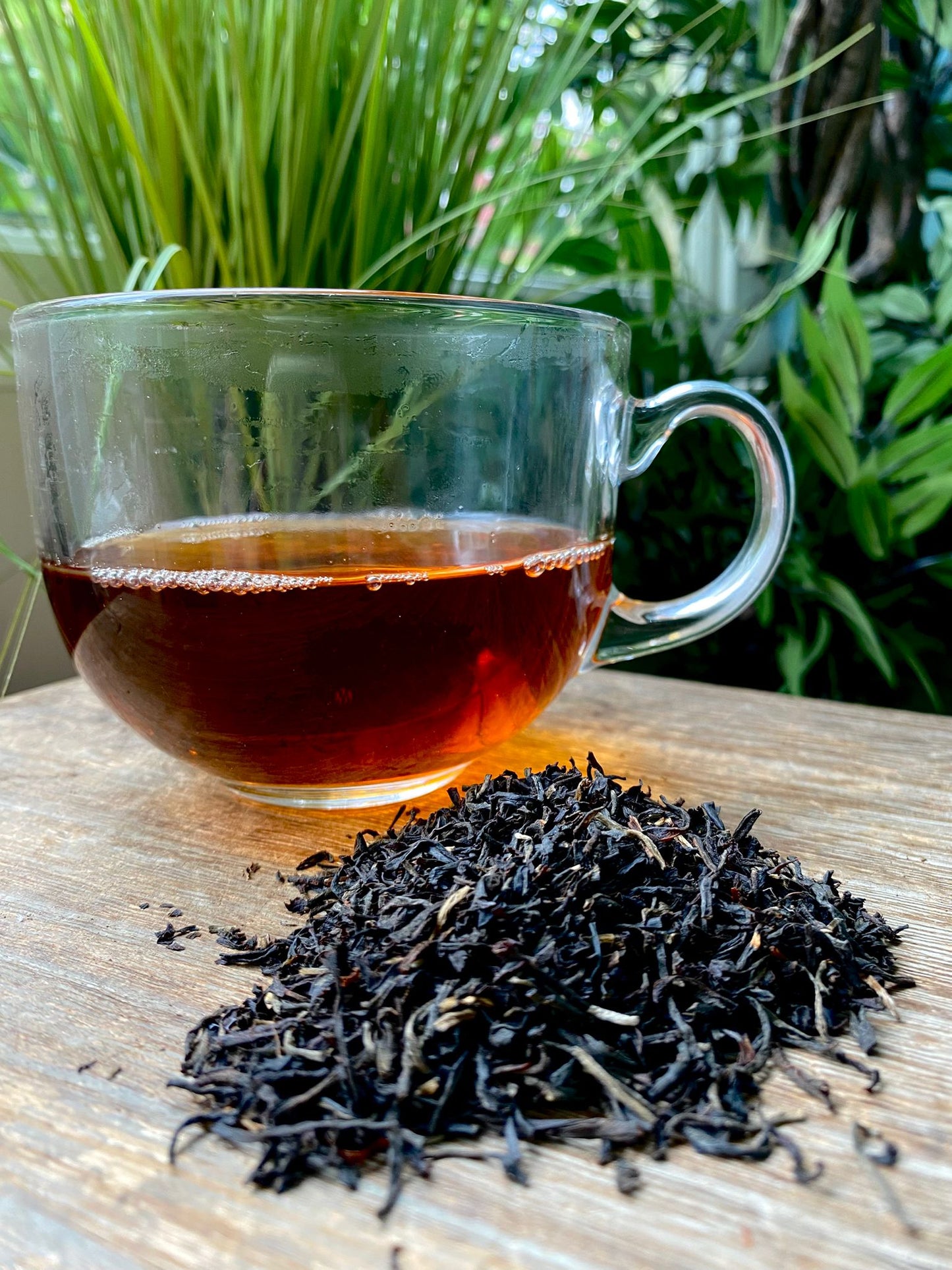 A clear glass cup of black tea sits on a wooden surface next to a pile of Assam loose leaf tea; green plants are in the background.