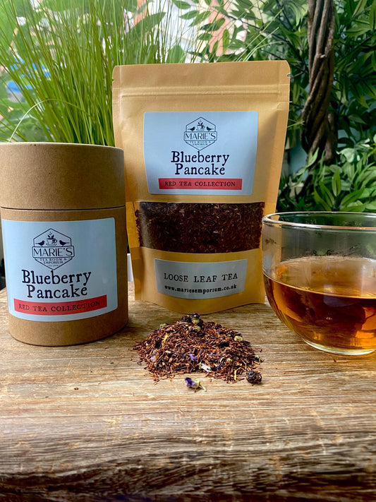 Maries Loose leaf tea labeled "Blueberry Pancake" is displayed on a wooden surface, with a glass of brewed tea nearby. Greenery in the background adds freshness.