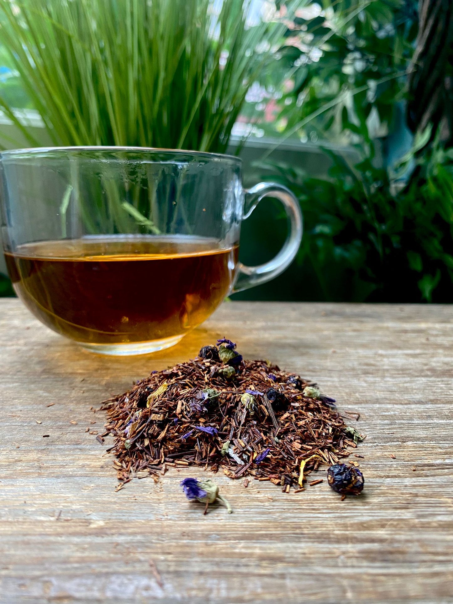 A clear glass cup of red tea sits on a wooden table near a pile of marie's blueberry pancake loose leaf tea.