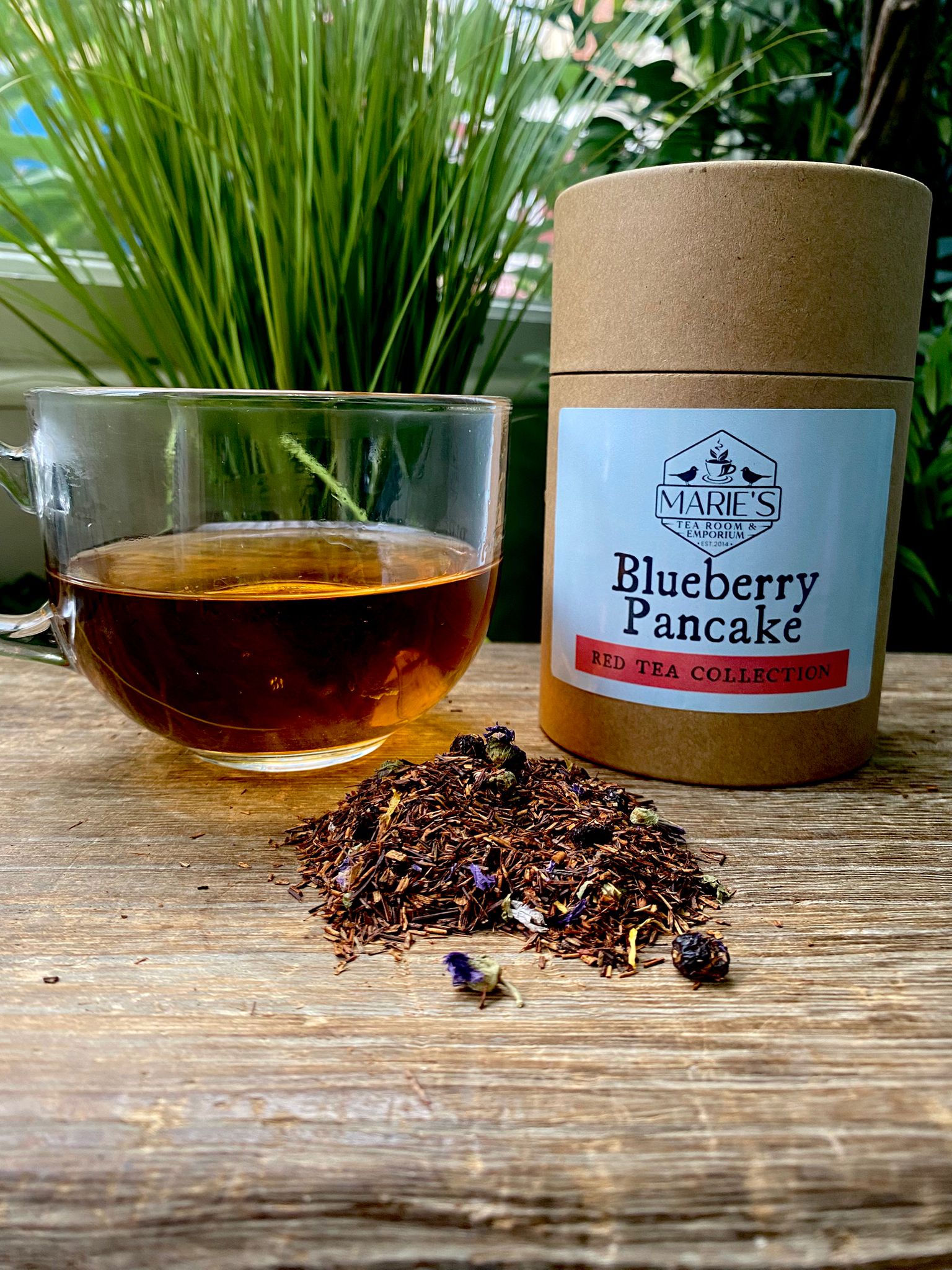 A glass cup of reddish-brown tea sits on a rustic wooden table beside a cylindrical tin labeled "MARIE'S Blueberry Pancake Red Tea Collection." Loose tea leaves and blueberries are scattered on the table, with lush green foliage in the background.