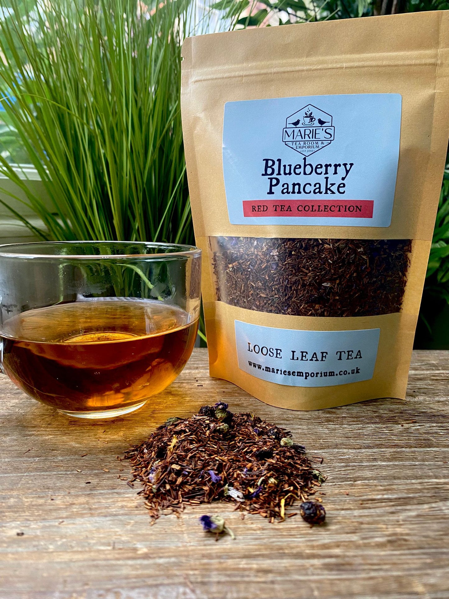 A glass cup of amber tea is on a wooden table beside a brown paper bag labeled "maries Blueberry Pancake, Red Tea Collection." Loose tea is scattered nearby.