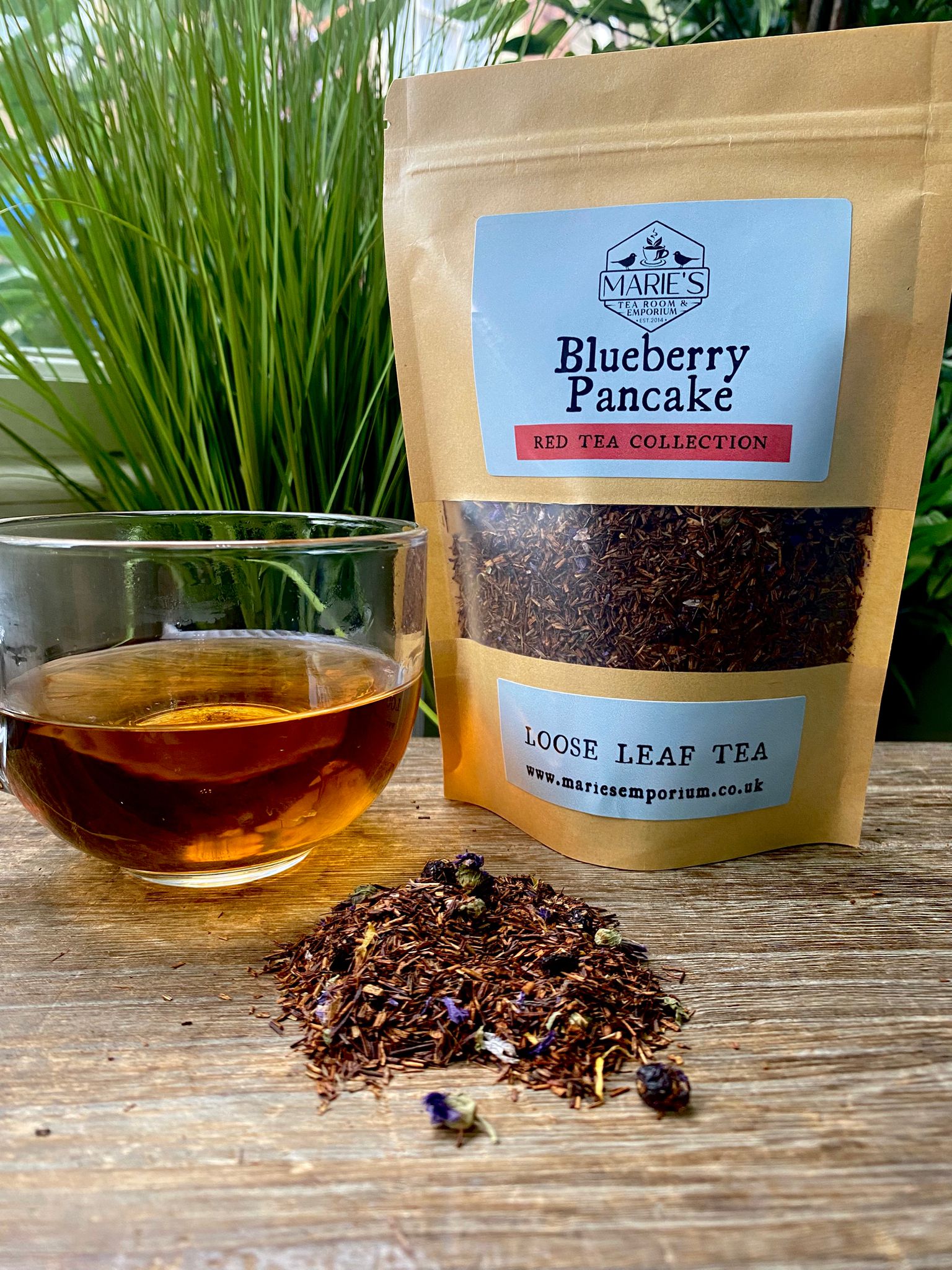 A glass cup of amber tea is on a wooden table beside a brown paper bag labeled "maries Blueberry Pancake, Red Tea Collection." Loose tea is scattered nearby.