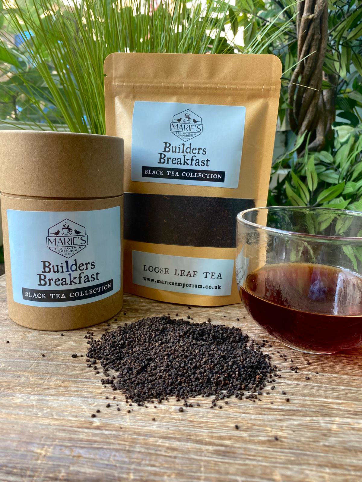 Two containers of 'Builders Breakfast' black tea with a glass of brewed tea on a wooden surface with greenery in the background.