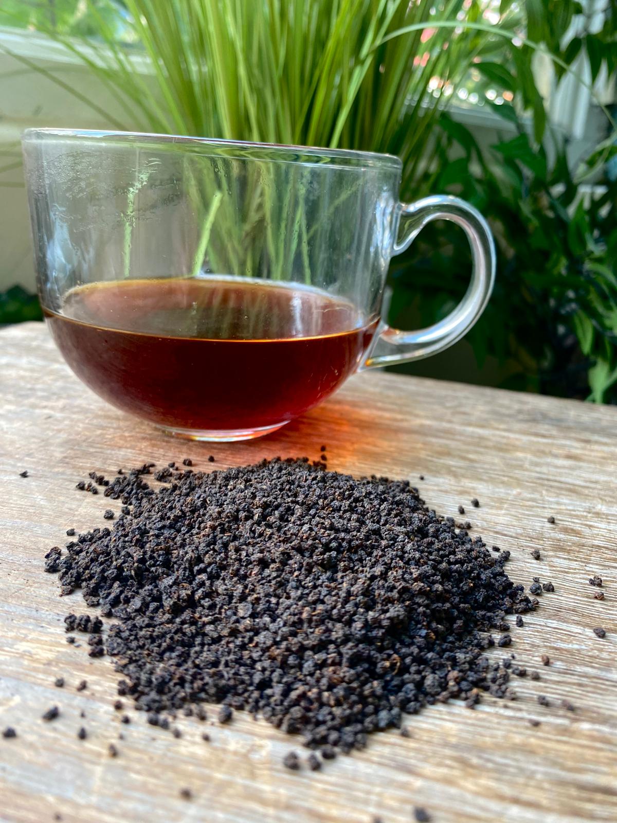 Clear glass mug with tea on a wooden surface with scattered tea leaves, green plants in the background