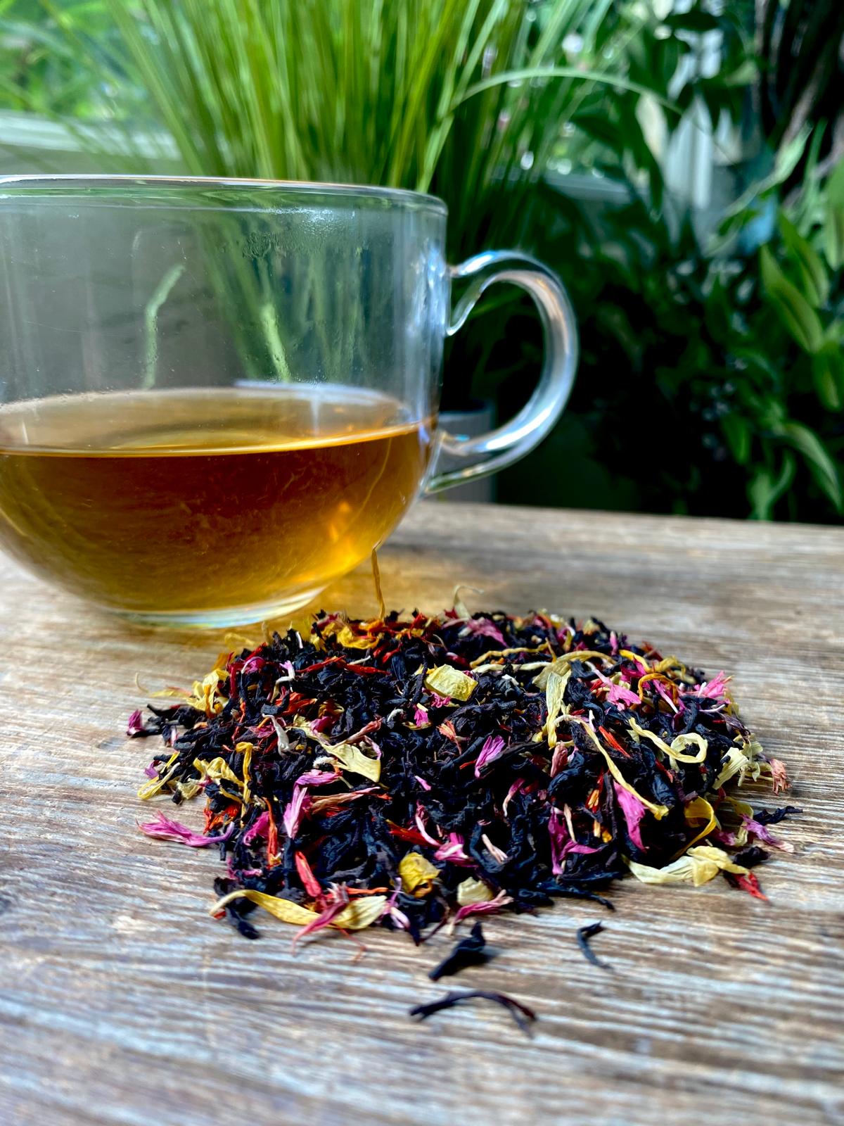 Clear glass mug of Campfire at Jamestown tea with a pile of dried tea leaves on a wooden surface with greenery in the background
