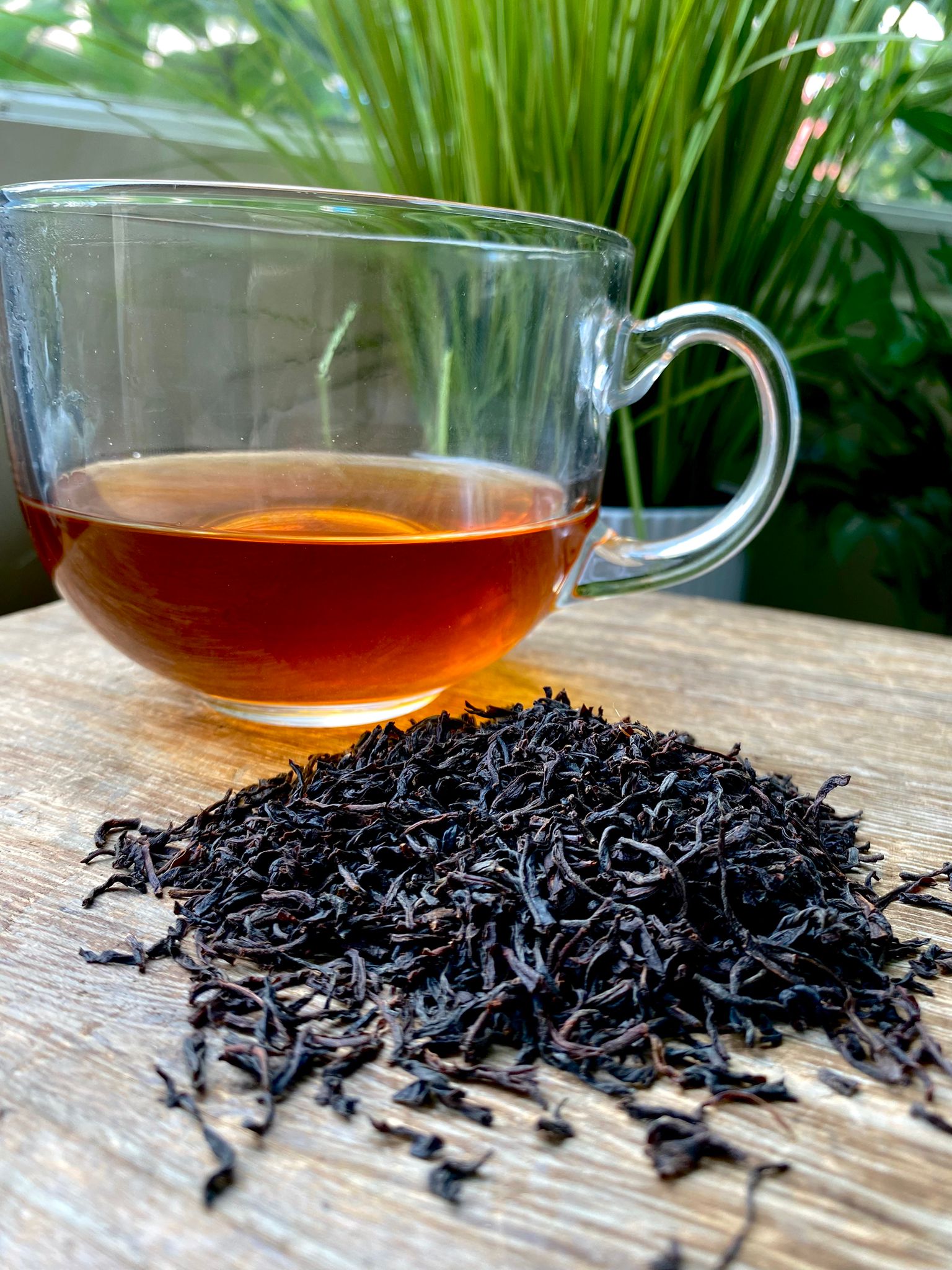 Clear glass mug with Ceylon tea and a pile of black tea leaves on a wooden surface with greenery in the background.