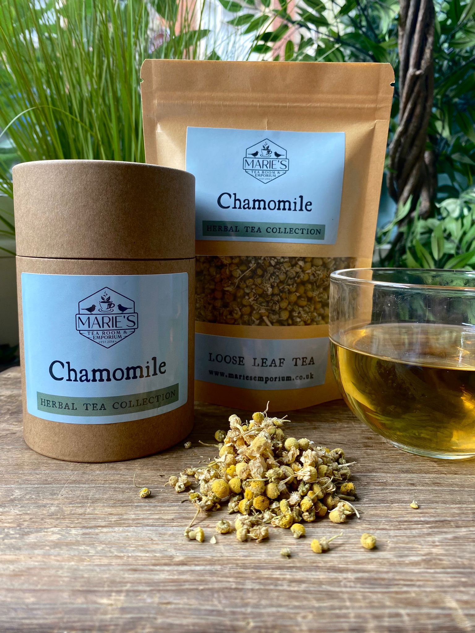 Marie's Loose Tea Collection with chamomile tea packaging, dried  loose leaf chamomile flowers, and a glass of tea on a wooden surface.