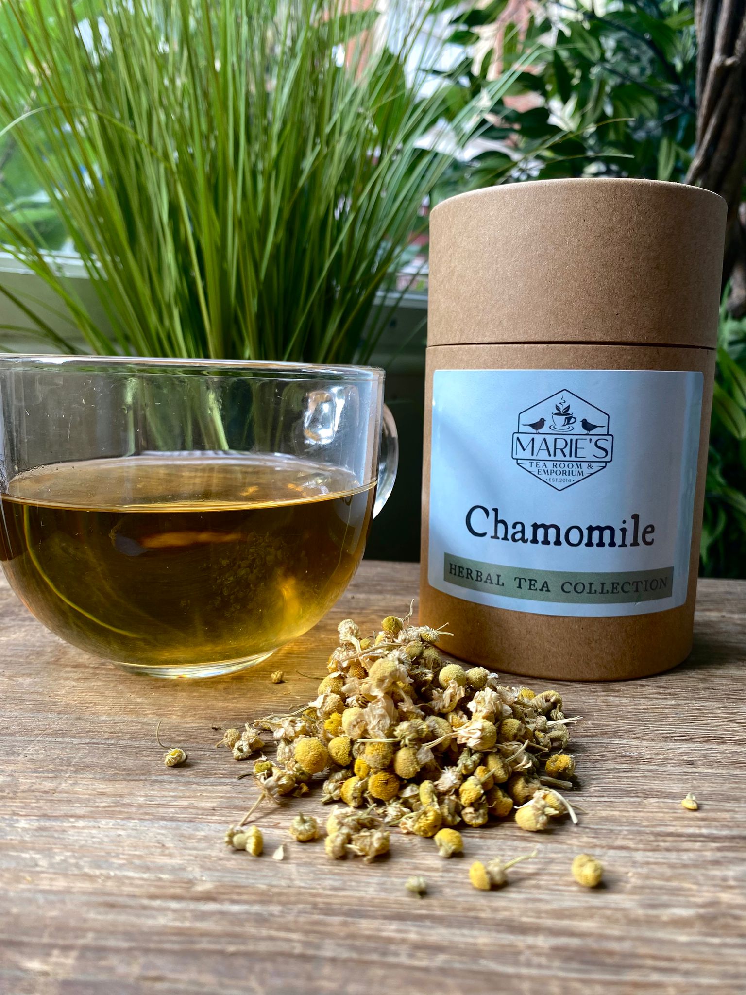 maries loose leaf Chamomile tea with a glass of tea and dried chamomile flowers on a wooden surface, with 'Marie's Tea' packaging in the background.