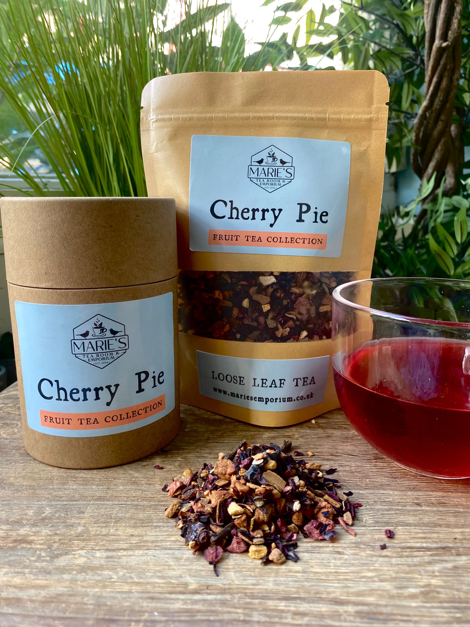 Marie's Tea Collection Cherry Pie tea packaging with loose tea leaves and a glass of loose leaf tea on a wooden surface.
