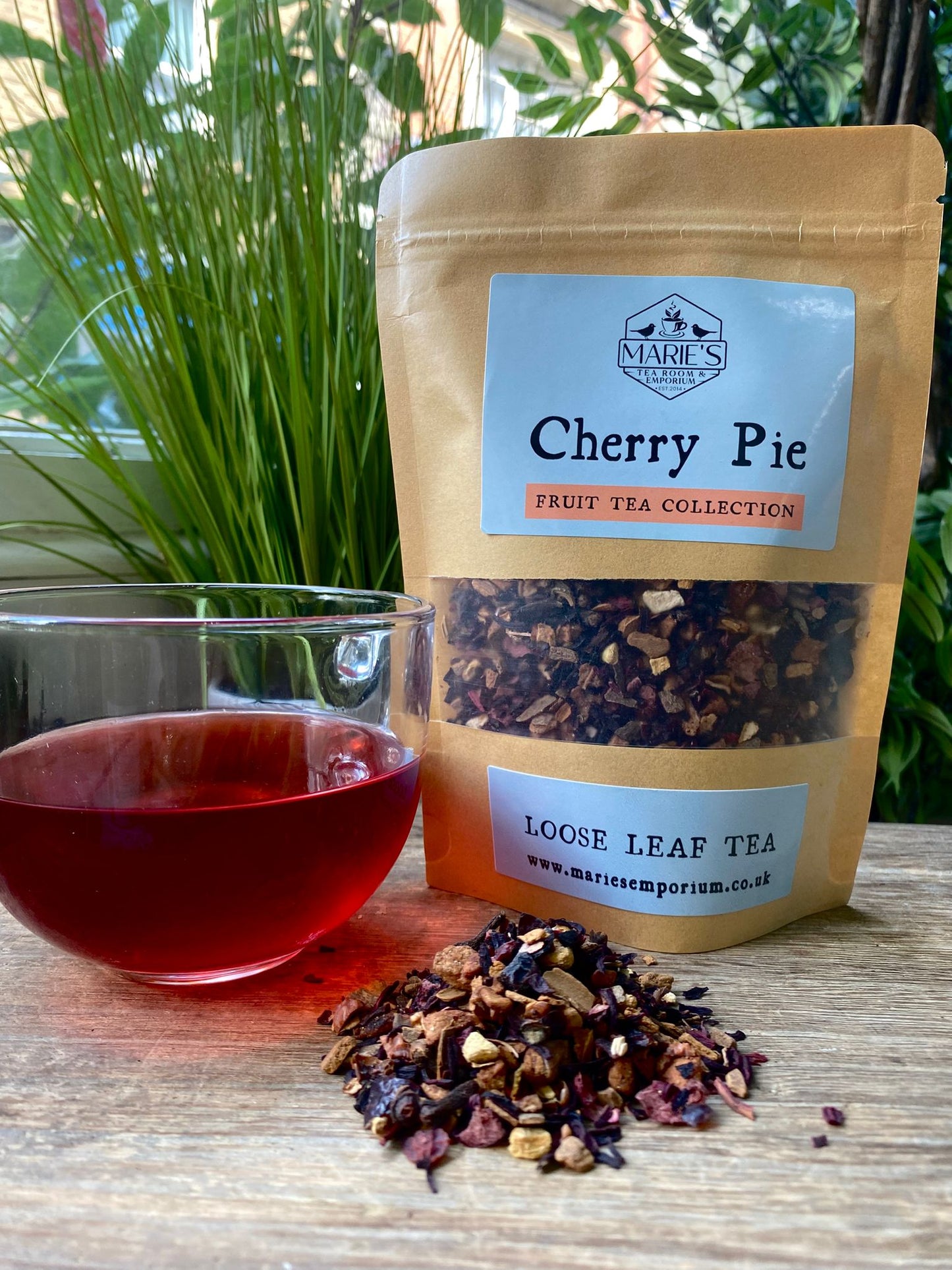 Maries Loose leaf tea package labeled 'Cherry Pie' from Marie's Emporium with a glass of brewed tea and scattered tea leaves on a wooden surface.