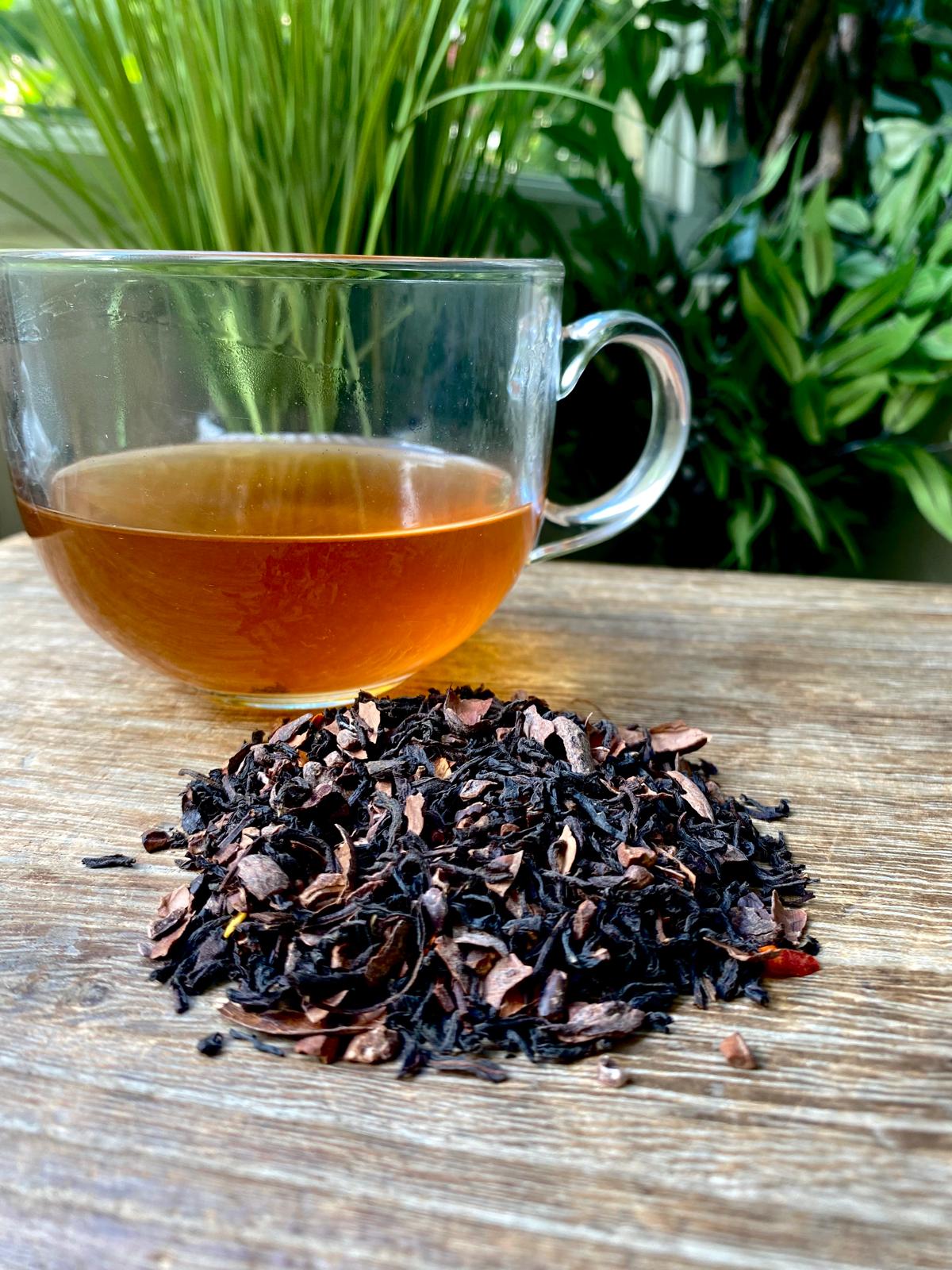 A glass mug of Chilli chocolate tea sits on a wooden surface beside a pile of loose black tea leaves, set against a backdrop of lush green plants.