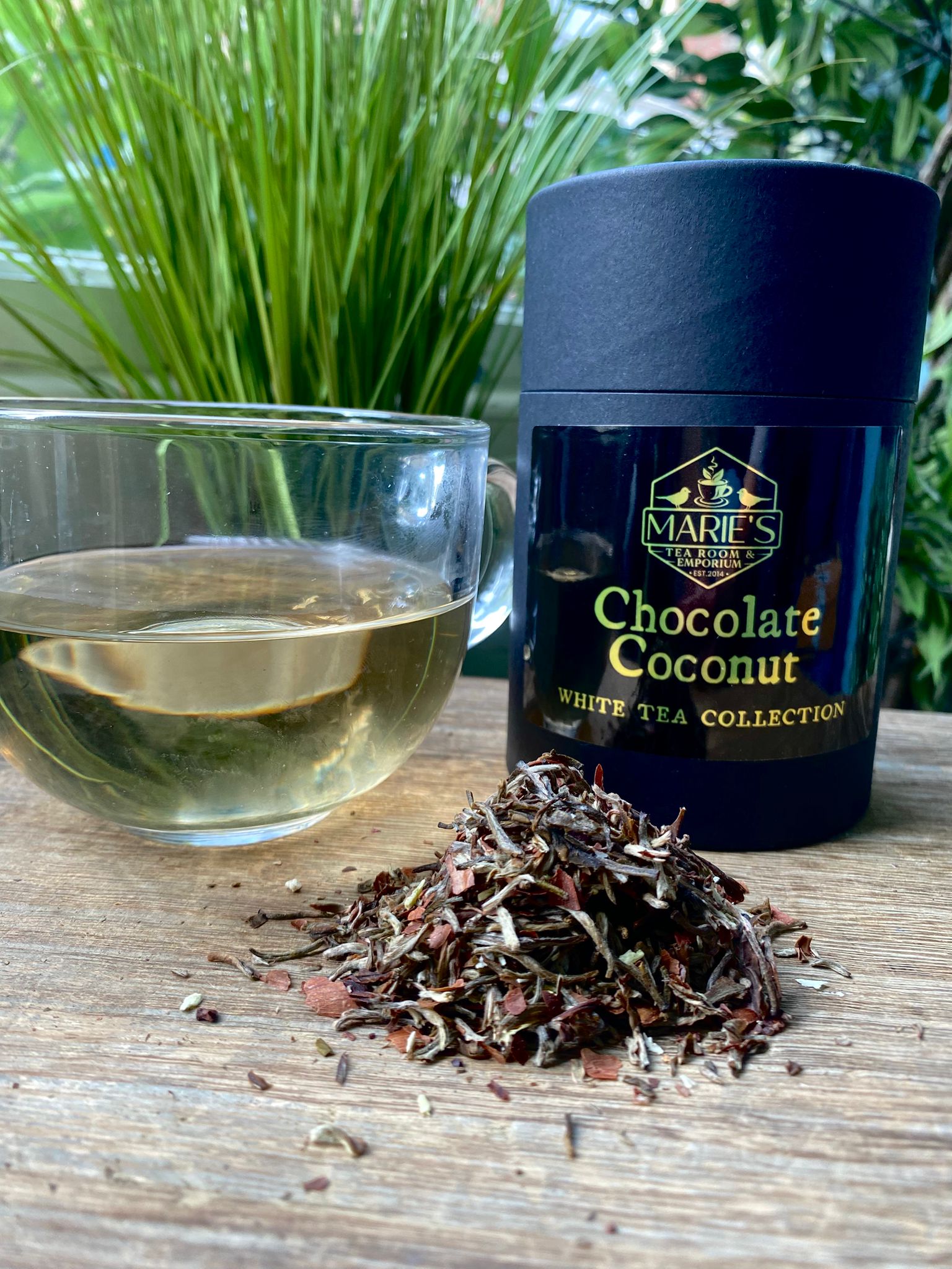 Jar of Marie's Chocolate Coconut White Tea Collection with a glass of tea and tea leaves on a wooden surface.