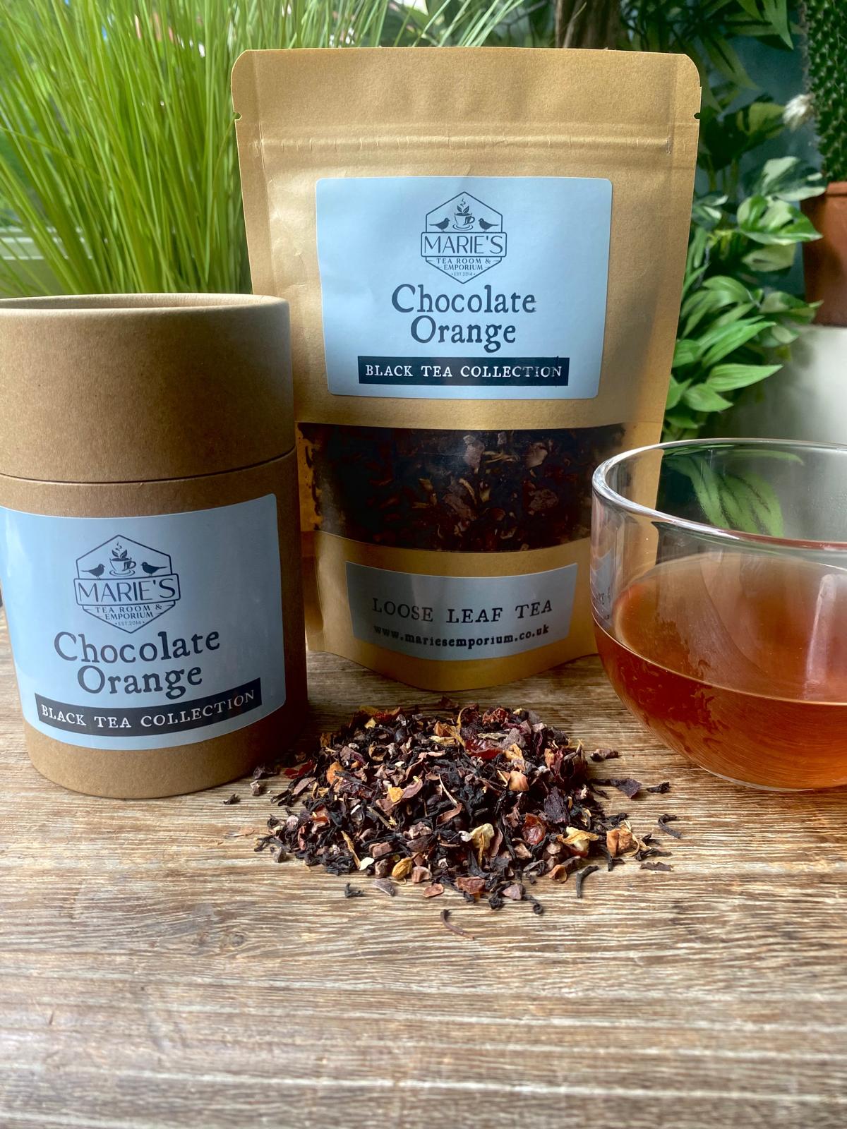 Containers and a pouch of Marie's Chocolate Orange black tea are on a wooden surface with a glass cup of brewed tea amidst fresh greenery.