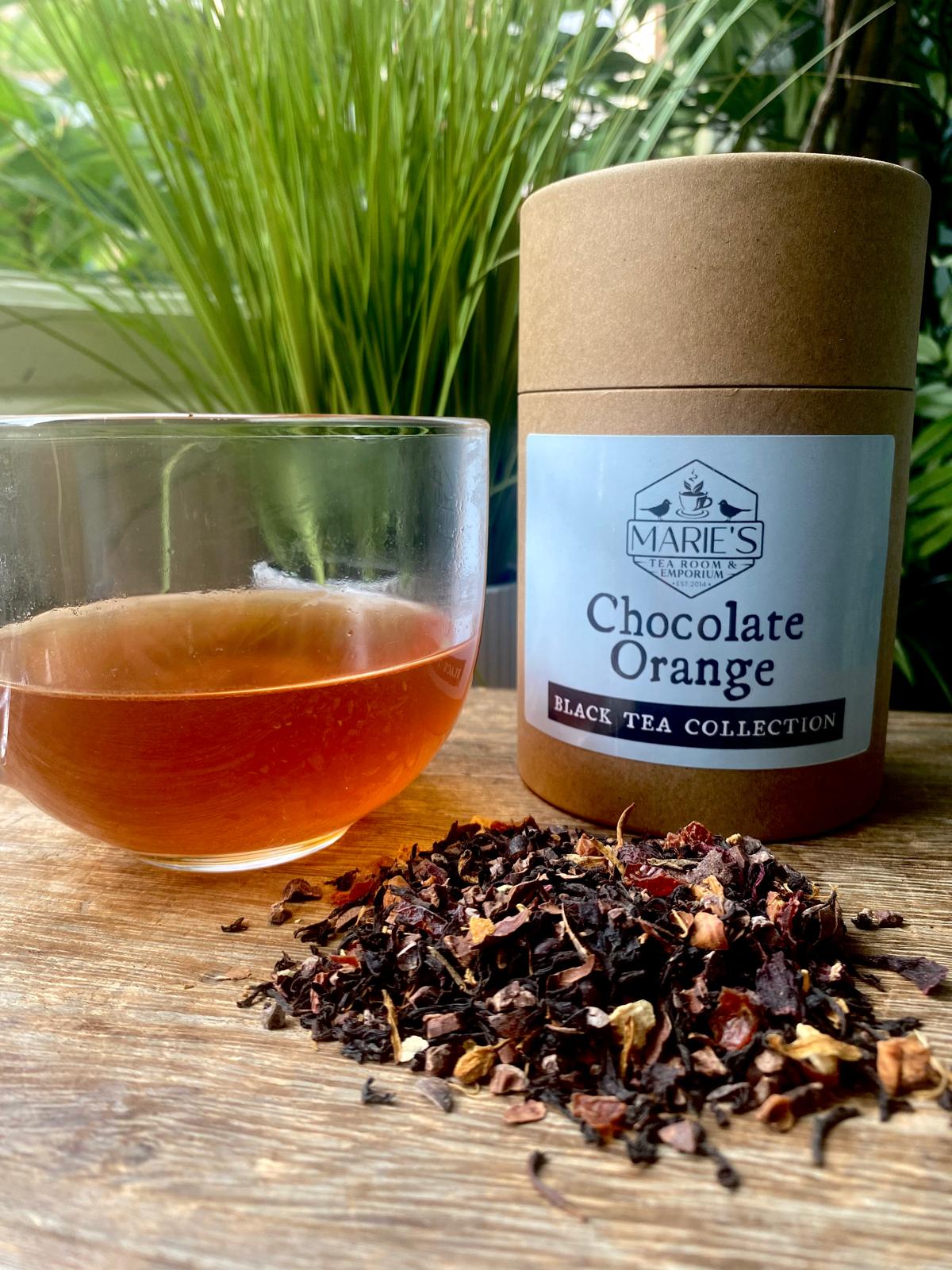 A glass of amber tea sits on a wooden surface beside a cylindrical container labeled "Chocolate Orange Black Tea Collection." Loose tea leaves are scattered, with lush greenery in the background. The scene conveys a fresh and inviting atmosphere.