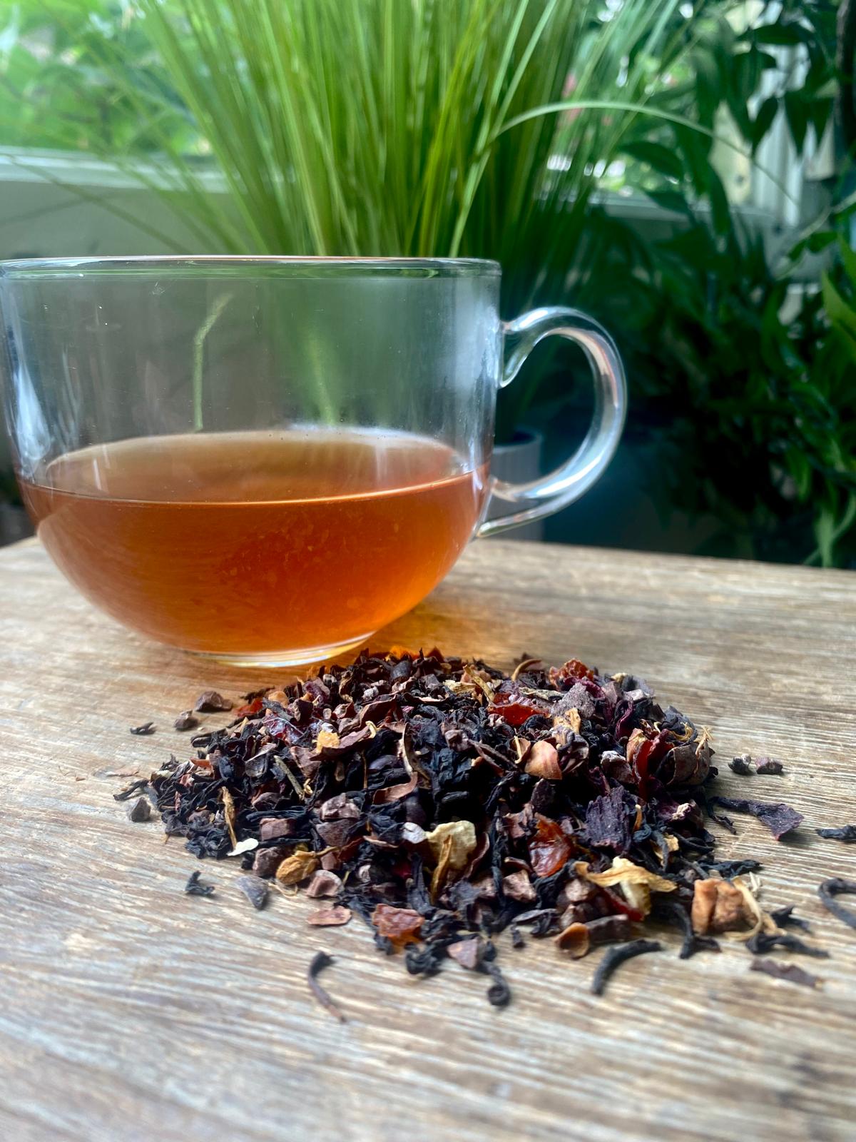 A clear glass cup filled with Chocolate orange tea sits on a wooden table beside a pile of dried tea leaves. Lush green plants are blurred in the background.