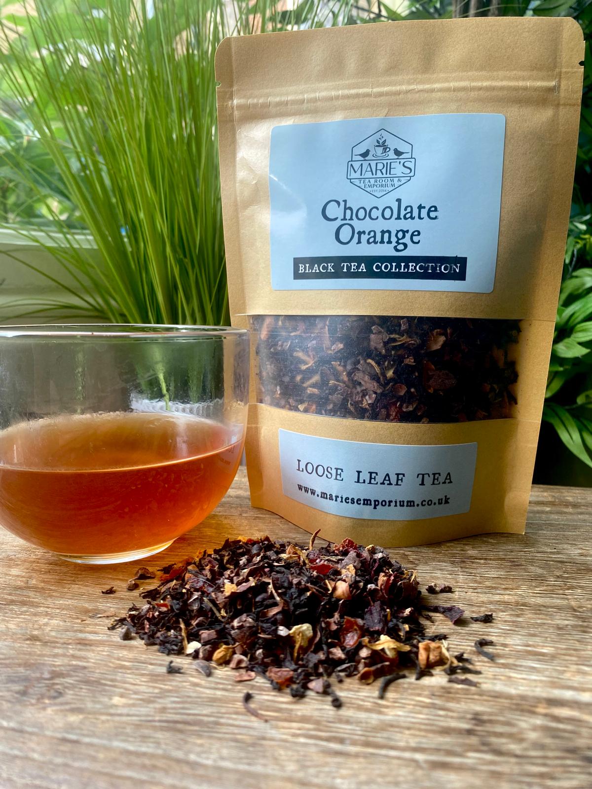 A clear glass cup filled with amber tea sits on a wooden table next to a pouch labeled "Chocolate Orange Black Tea." Loose leaf tea is scattered in the foreground with lush green plants in the background. The scene is warm and inviting.