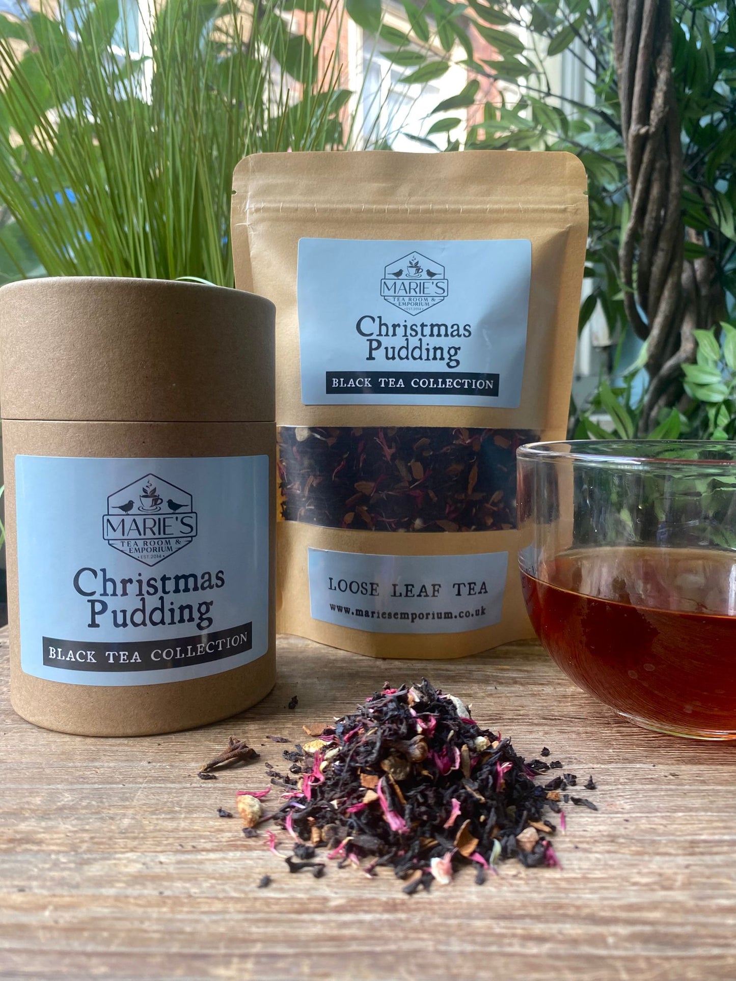 Two containers of Marie's Tea Company Christmas Pudding black tea, a glass of tea, and loose tea leaves on a wooden surface with plants in the background.