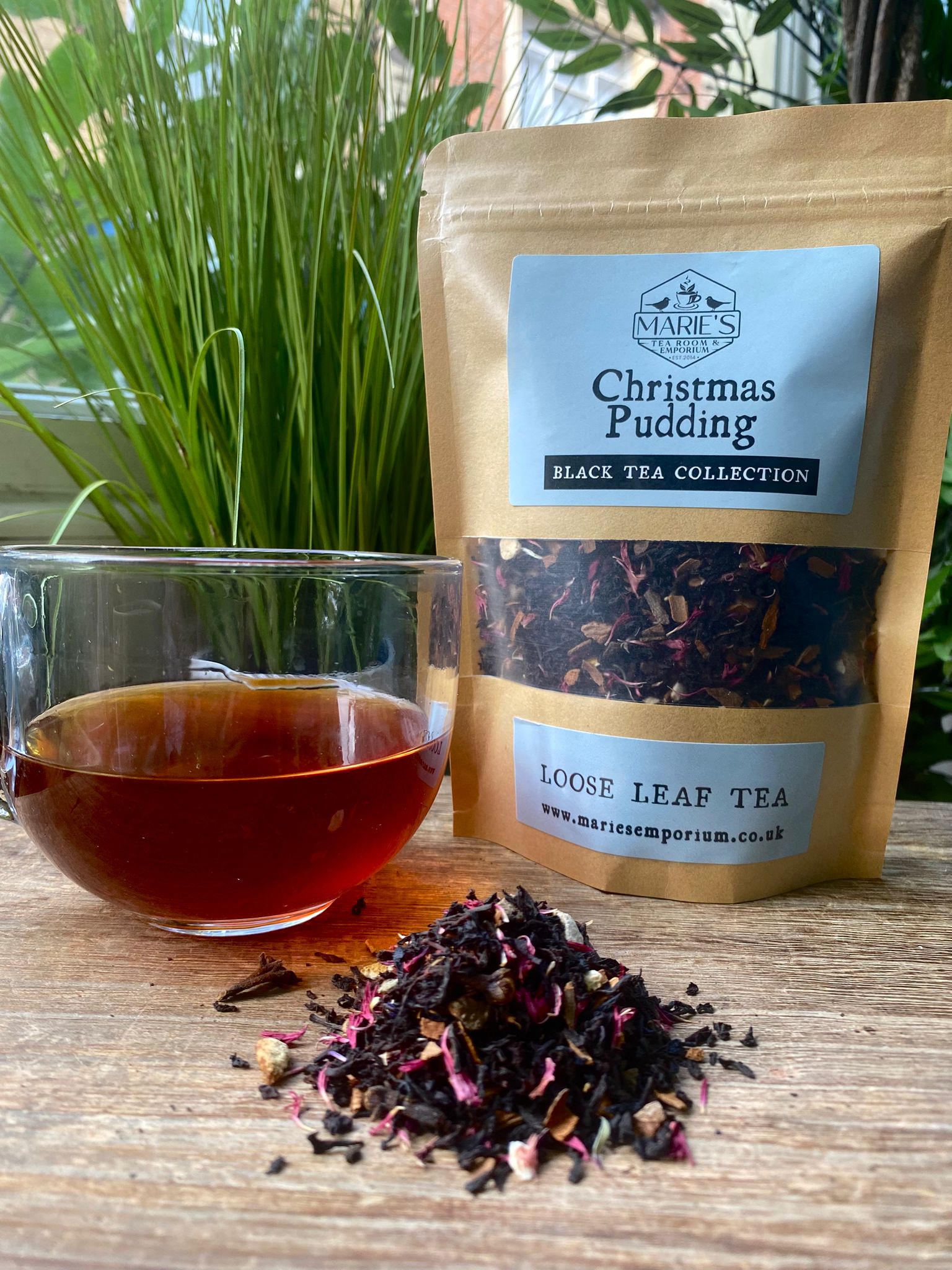 Tea packaging with a glass of tea and loose leaf tea on a wooden surface, with plants in the background.