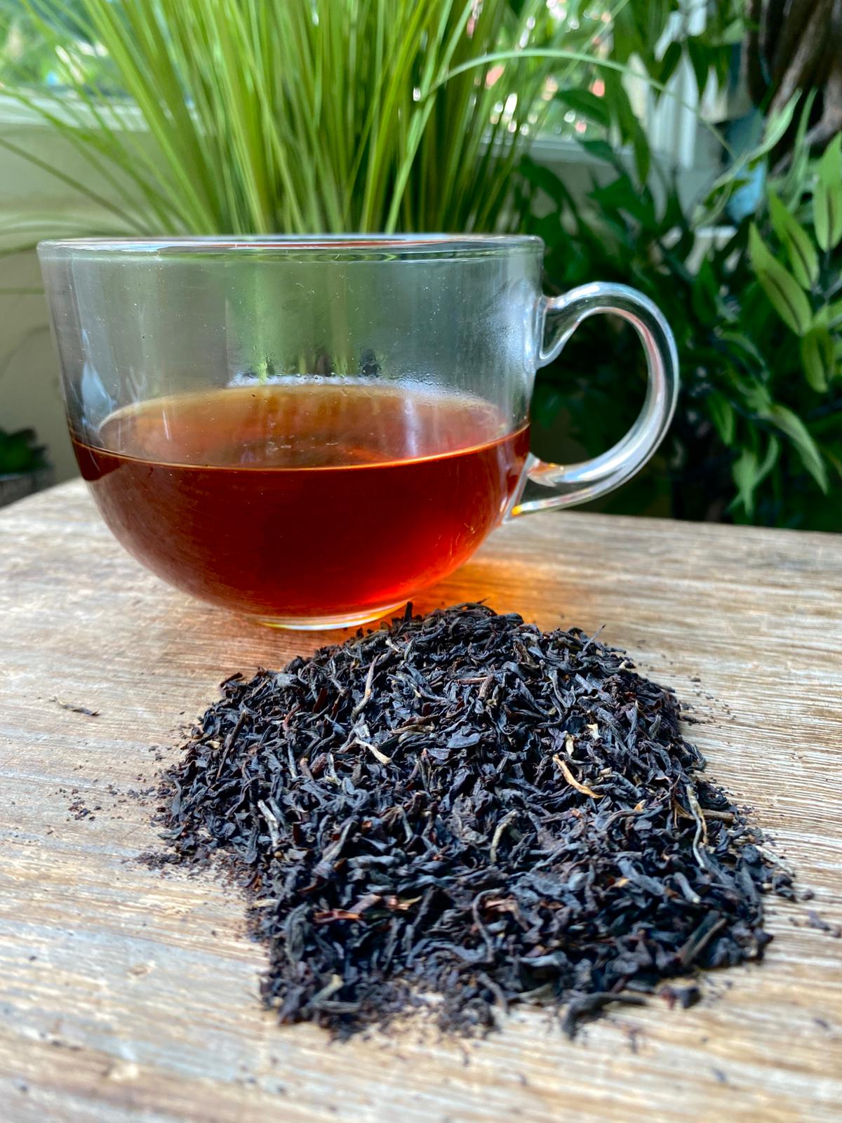 A glass cup of rich english breakfast tea sits on a wooden surface, next to a pile of loose black tea leaves. Background features lush green plants, conveying a serene, natural ambiance.