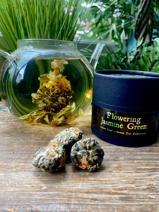 A glass teapot with blooming jasmine tea is next to a black container labeled "Flowering Jasmine Green." Three tea balls of loose leaf tea are in the foreground on a wooden table. Lush green foliage in the background.