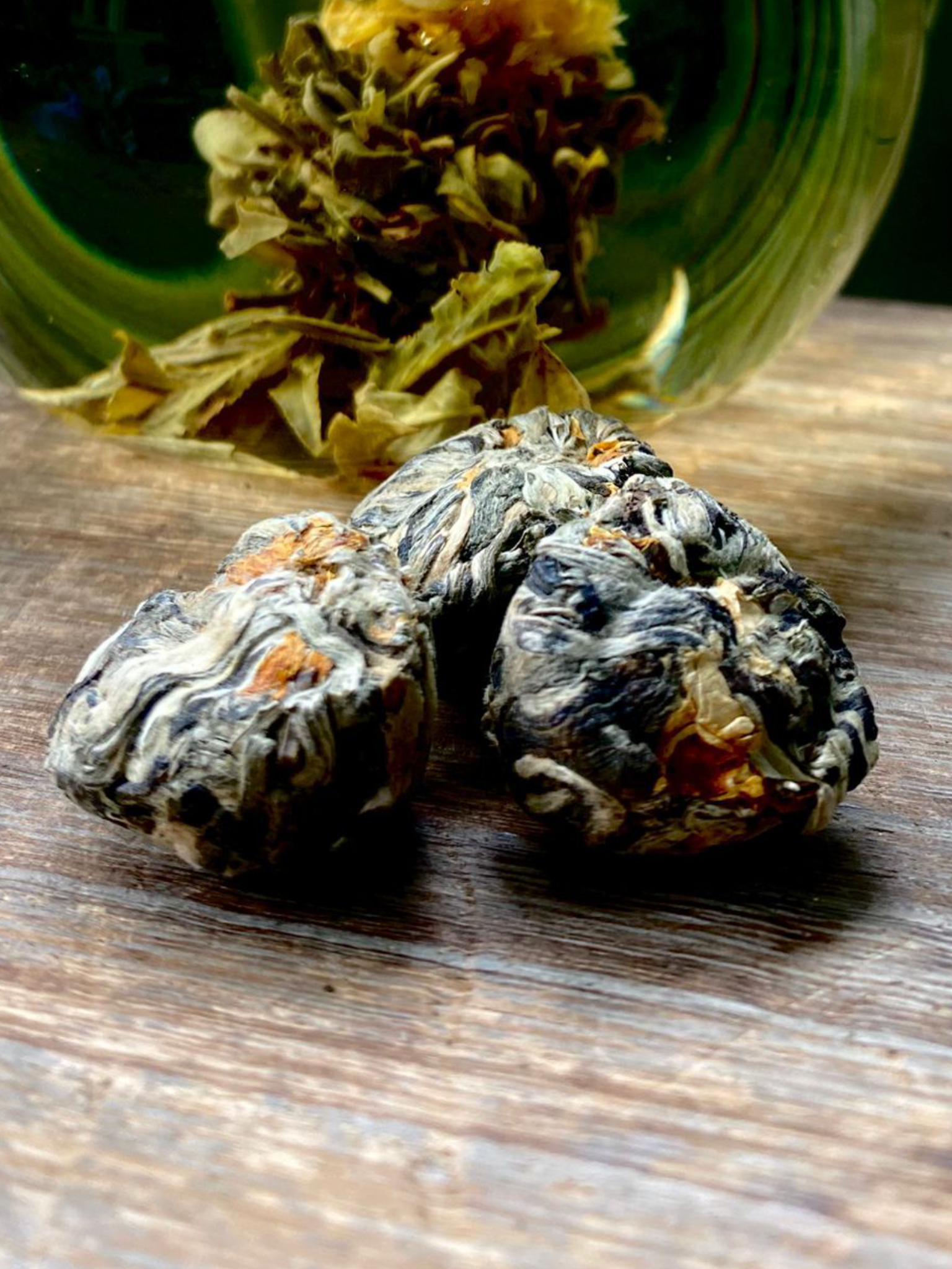 Three tightly rolled Flowering Jasmine loose leaf tea buds sit on a wooden surface, with a glass teapot in the background showing a blooming flower tea steeping.