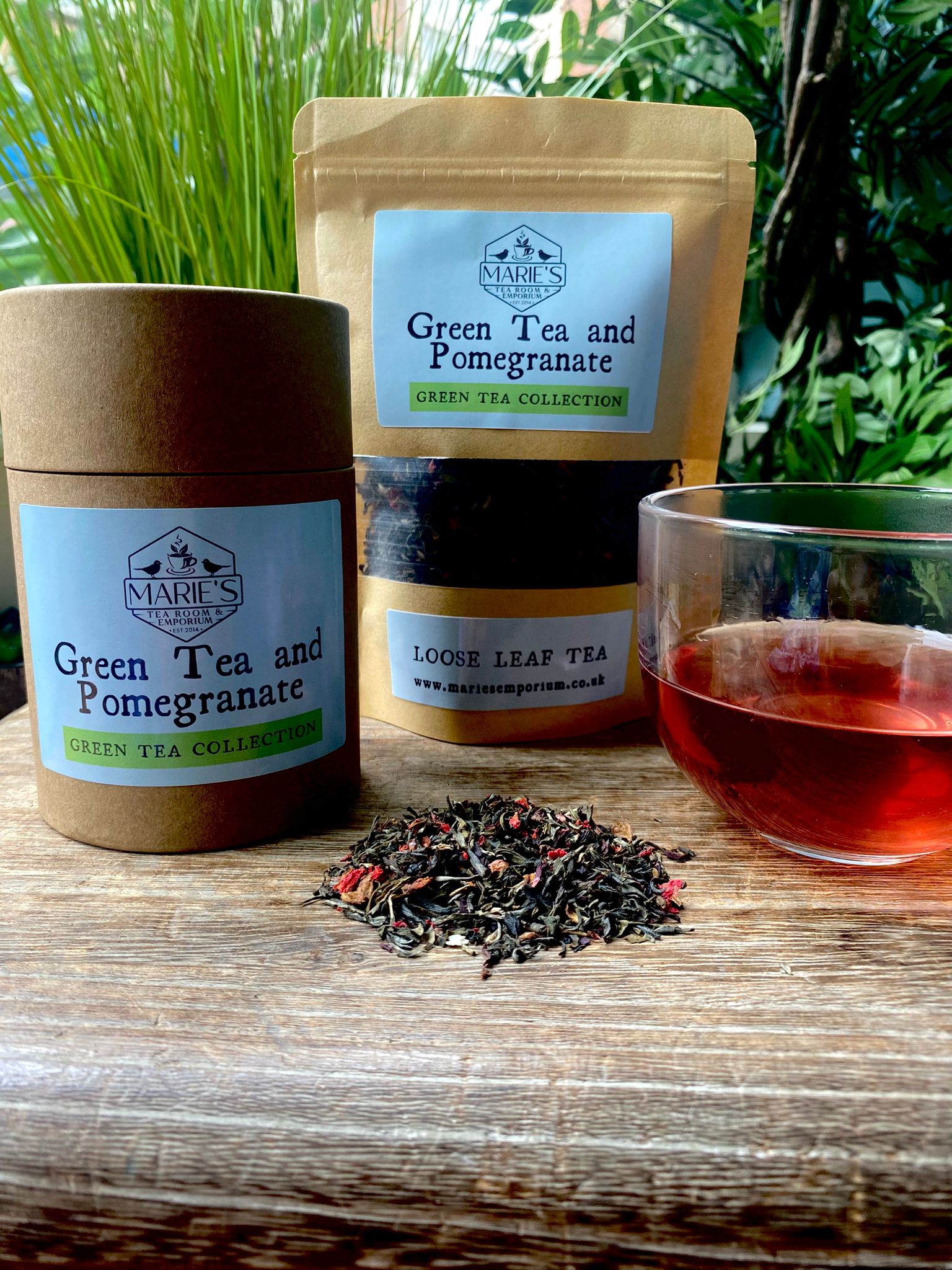 Maries Green Tea and Pomegranate tea packaging with a cup of tea on a wooden surface with scattered loose leaf tea leaves.