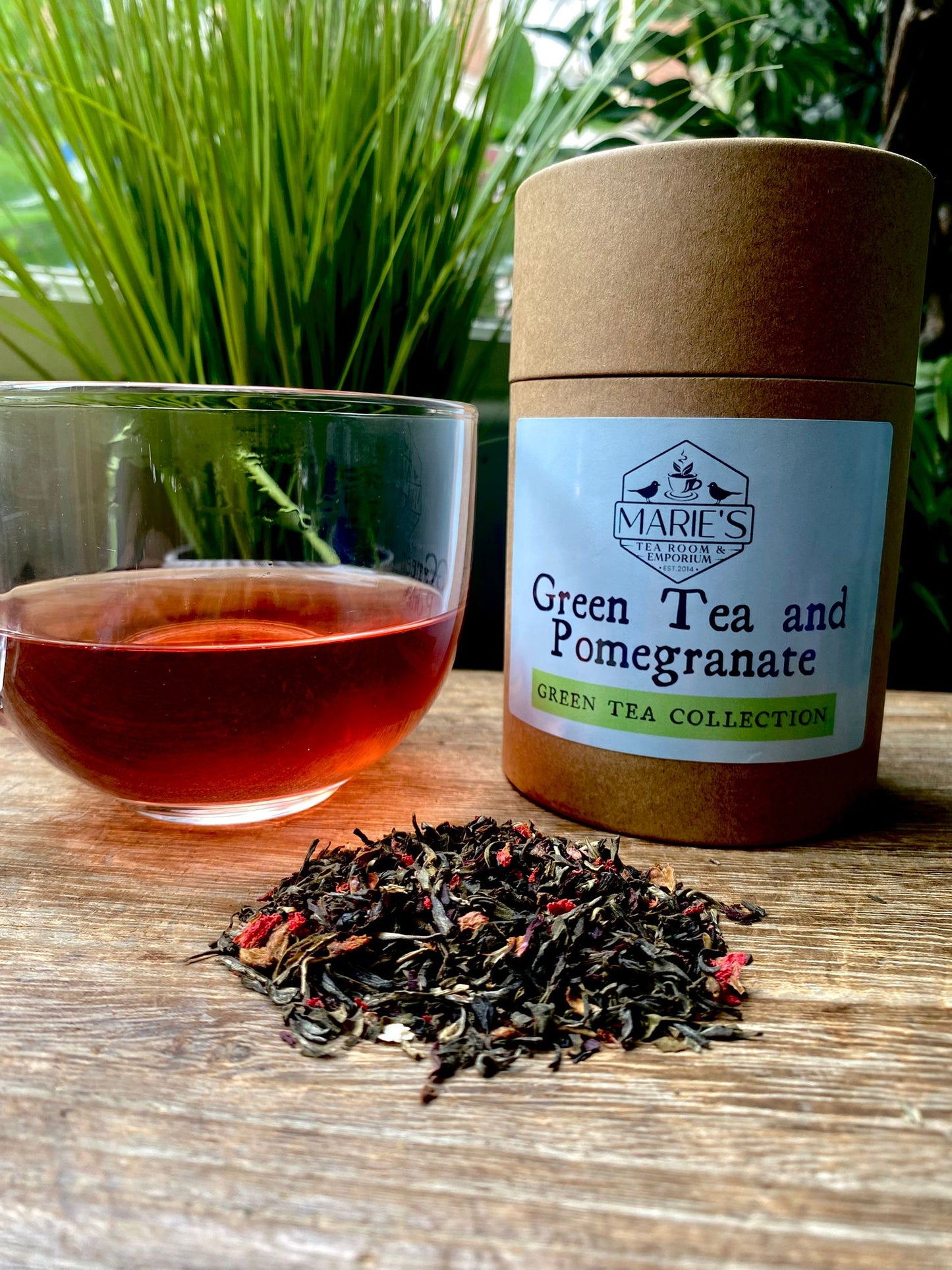 Green tea and pomegranate paper tin on a wooden surface with a glass of tea and loose leaf tea leaves.
