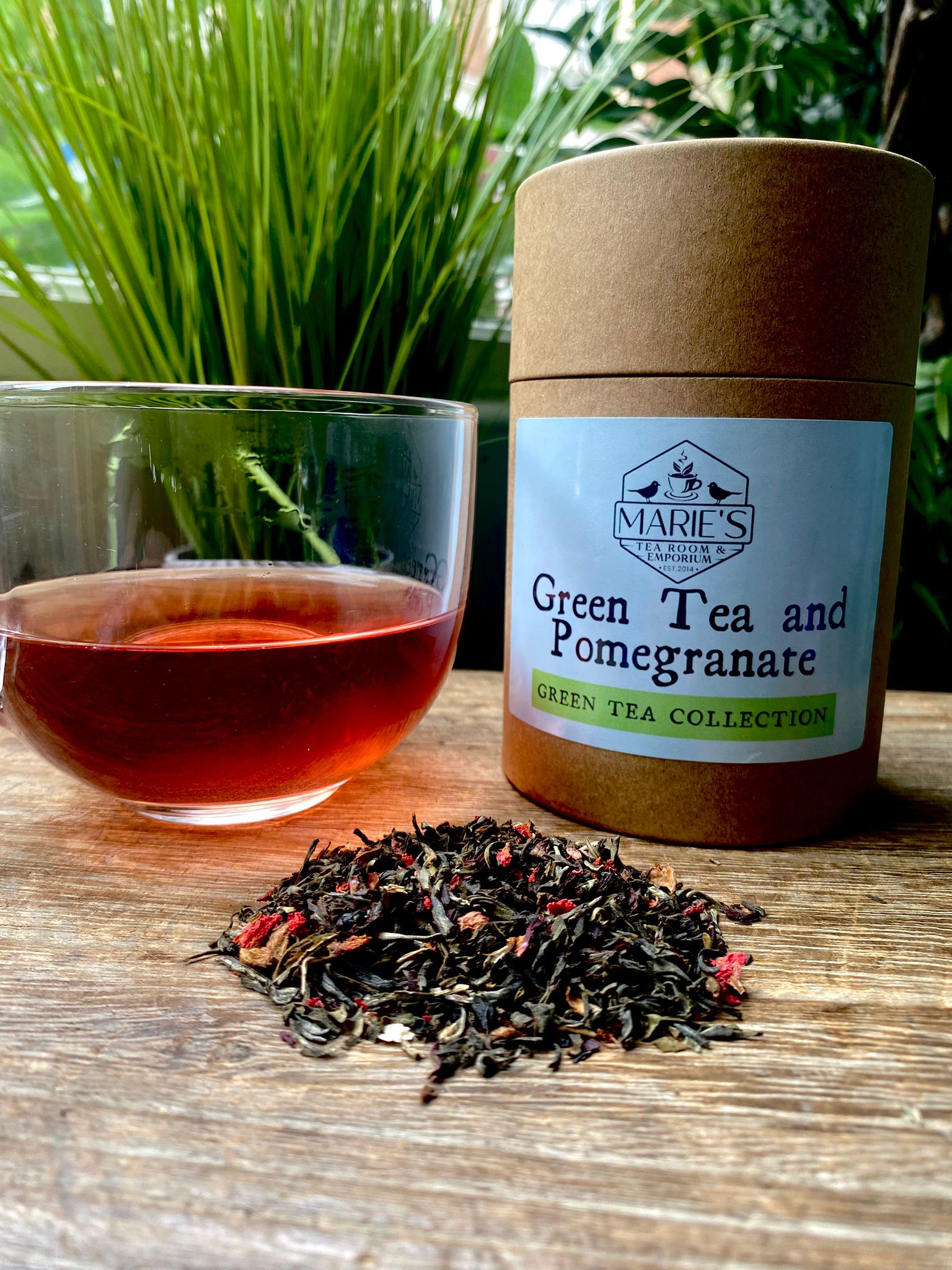 Green tea and pomegranate paper tin on a wooden surface with a glass of tea and loose leaf tea leaves.