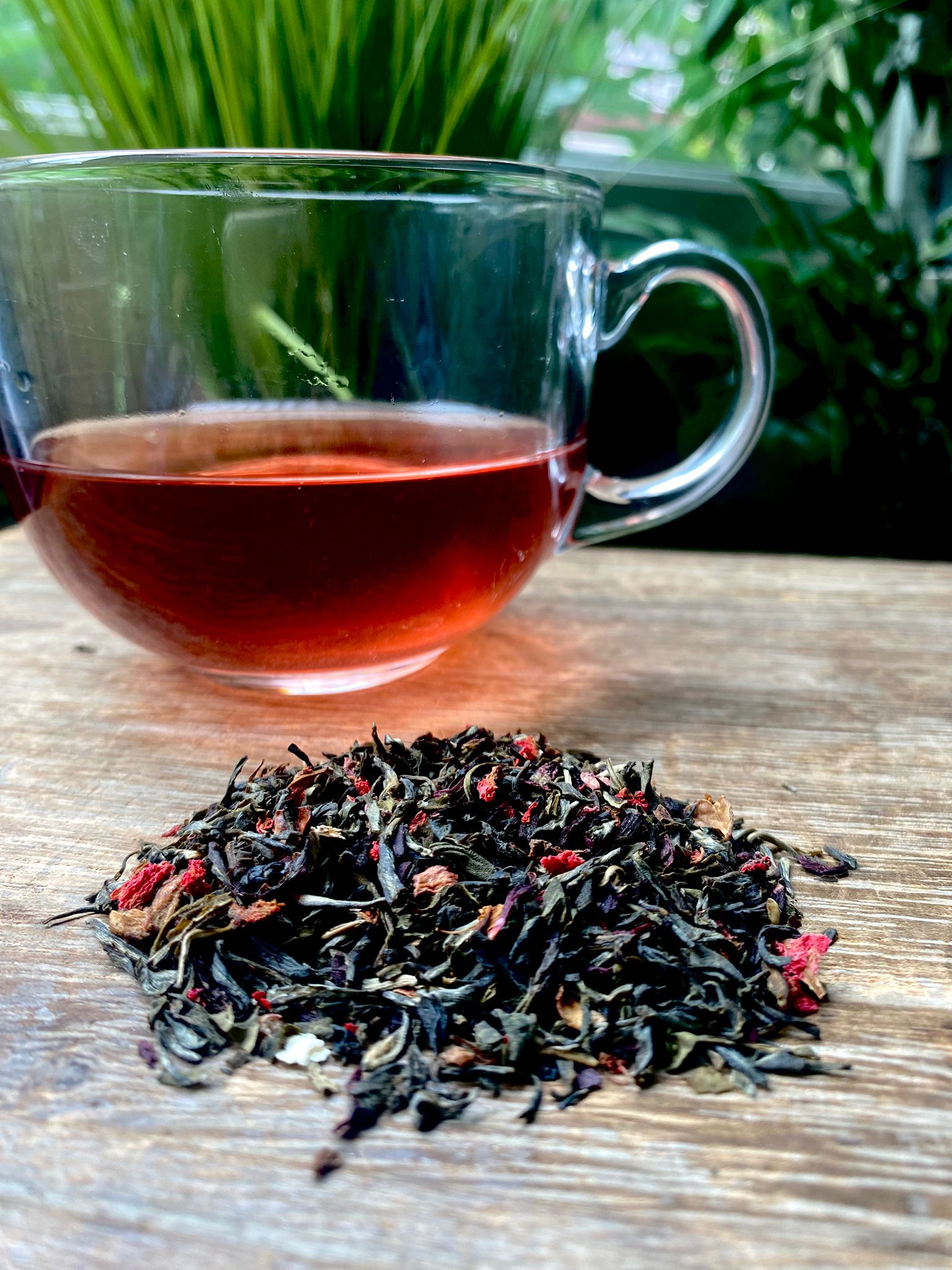 Clear glass mug with Green Tea and Pomegranate tea and a pile of green tea leaves on a wooden surface with greenery in the background.