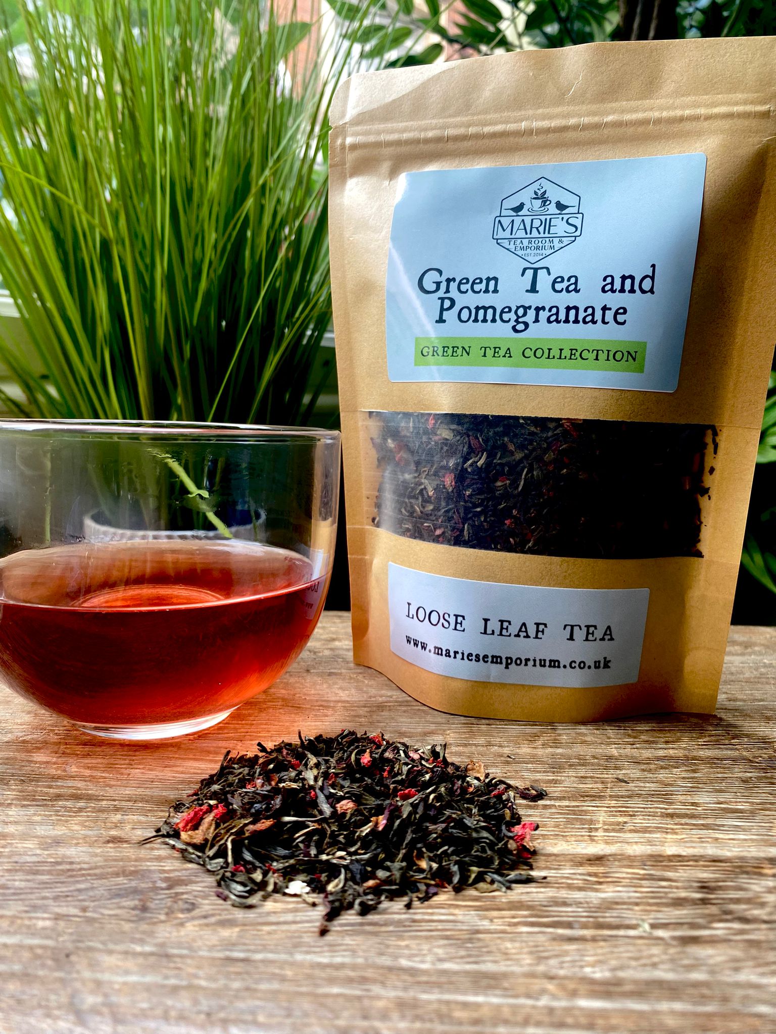 marie's Green Tea and Pomegranate tea package with a glass of tea and loose tea leaves on a wooden surface.