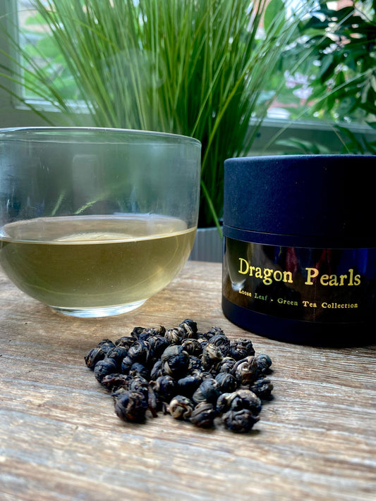 Tea leaves, a glass of tea, and a container labeled Marie's 'Dragon Pearls' on a wooden surface with plants in the background.