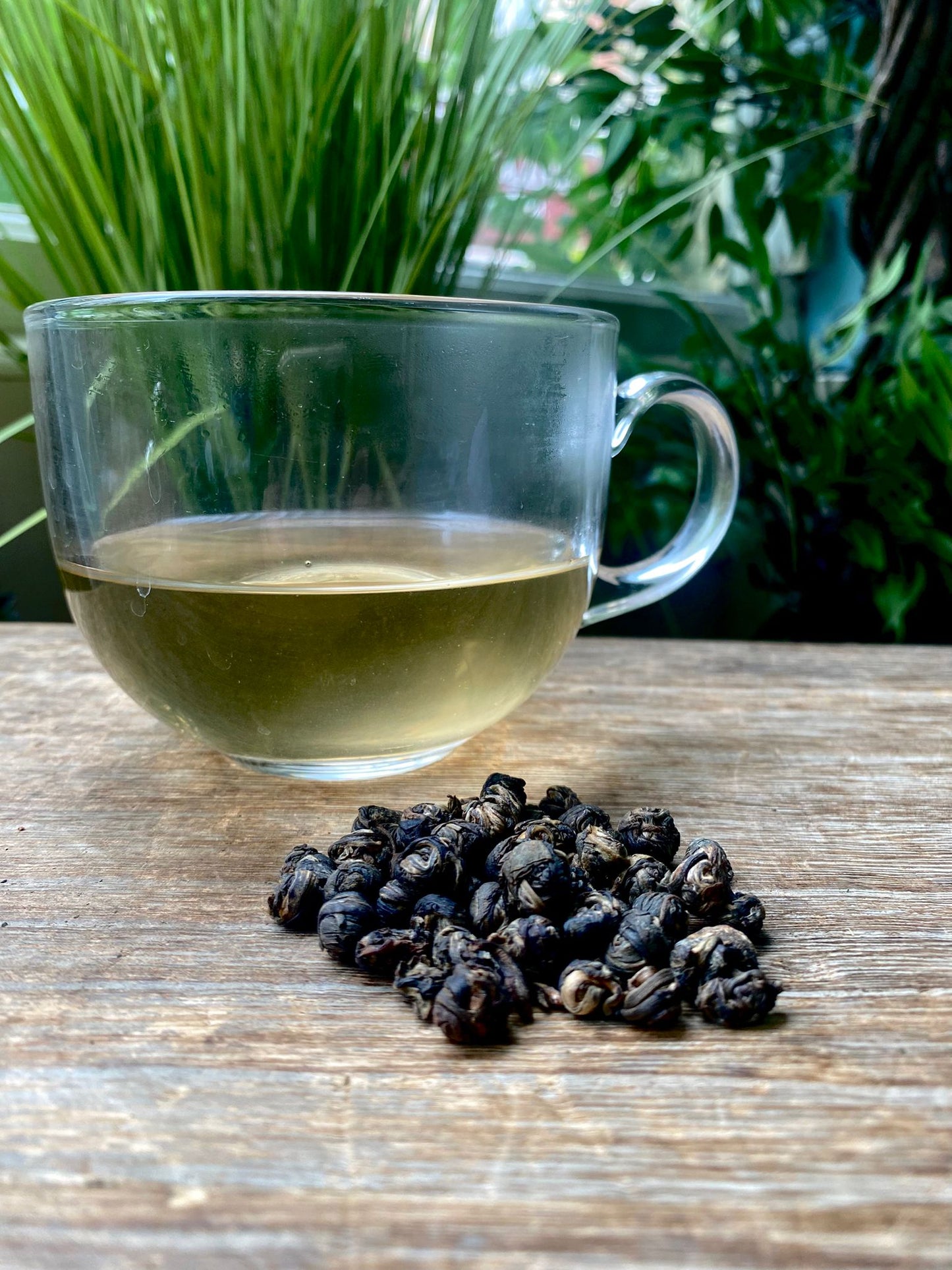 Clear glass mug of Jasmine dragon peals tea with loose leaf tea leaves on a wooden surface with greenery in the background