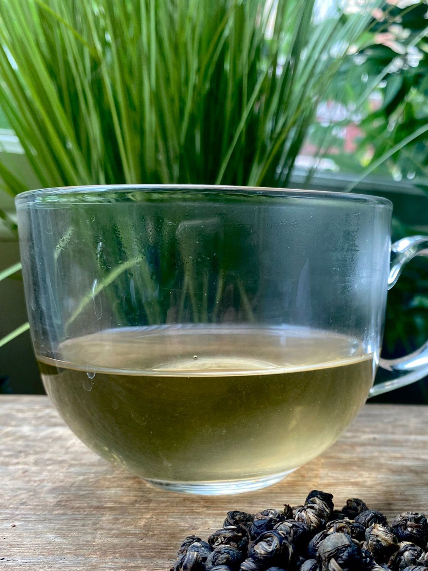 Clear glass mug of Jasmine dragon peals tea with loose leaf tea leaves on a wooden surface with greenery in the background