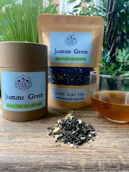 Loose jasmine green tea is displayed with a cardboard canister and a pouch labeled “Marie's Jasmine Green Tea.” A clear glass cup holds brewed tea. Verdant plants sit in the background.
