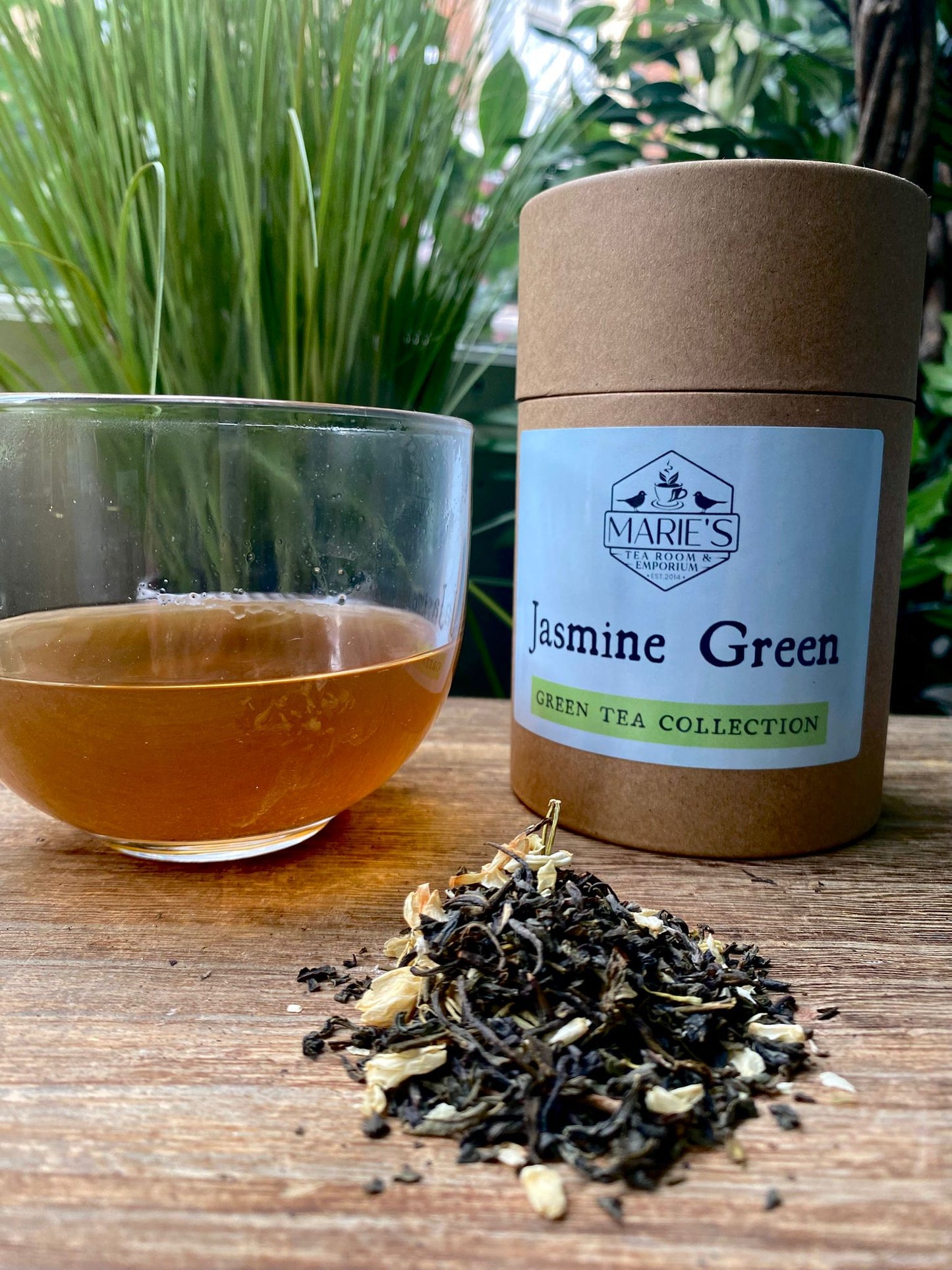 Marie's Jasmine Green tea packaging with a glass of tea and loose tea leaves on a wooden surface.