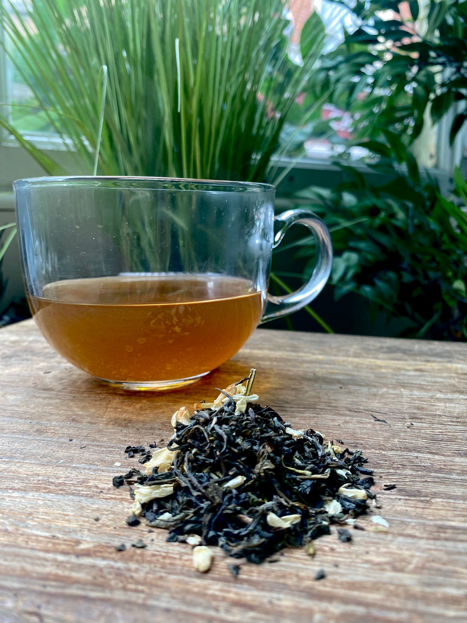 A glass cup of maries loose leaf jasmine green loose leaf tea sits on a wooden surface beside a small pile of loose leaf tea.