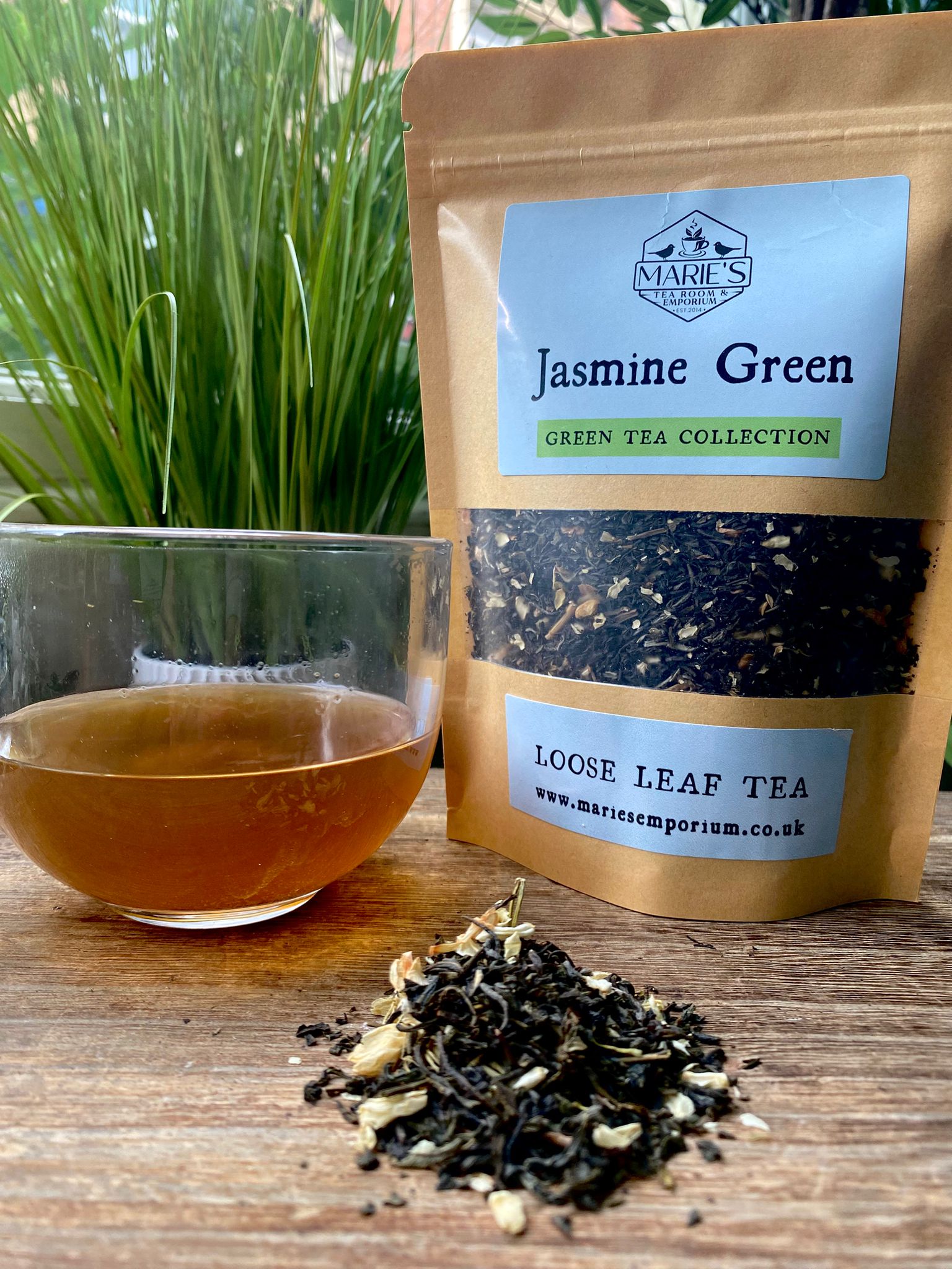 A clear glass cup filled with light brown maries jasmine green loose leaf tea tea sits beside a kraft paper bag of loose leaf tea, on a wooden table with green plants.